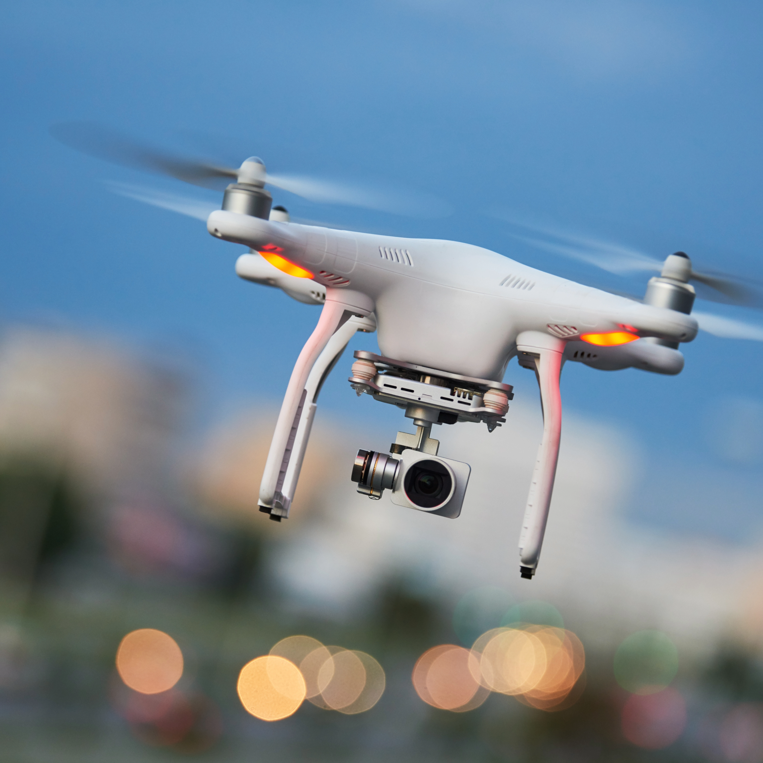 Quadcopter drone with camera hovering in the sky, with city lights blurred in the background.