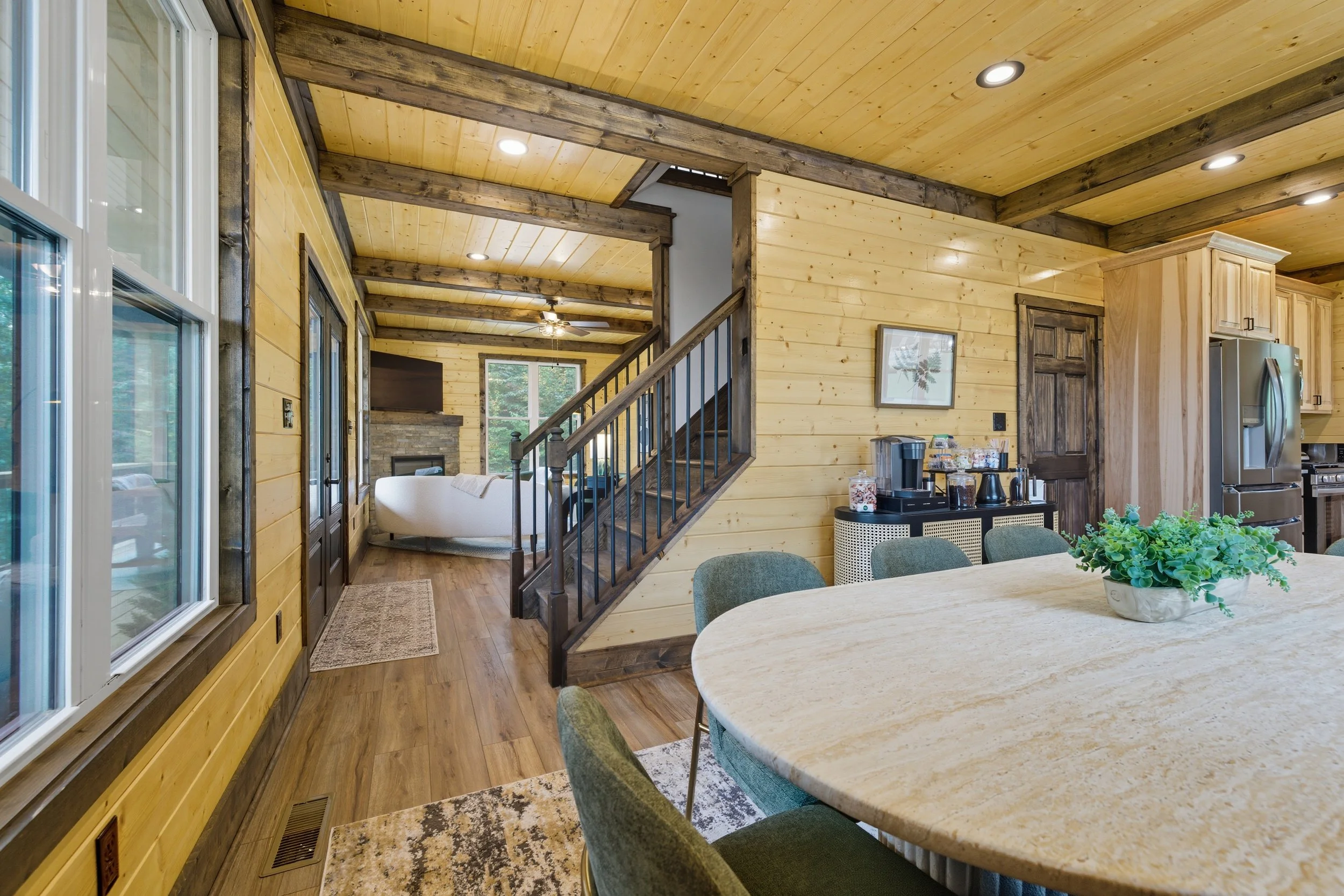 Interior of a wooden house with a kitchen and living area featuring wood-paneled walls, a coffee table, a white sofa, a television, and sliding glass doors leading outside.