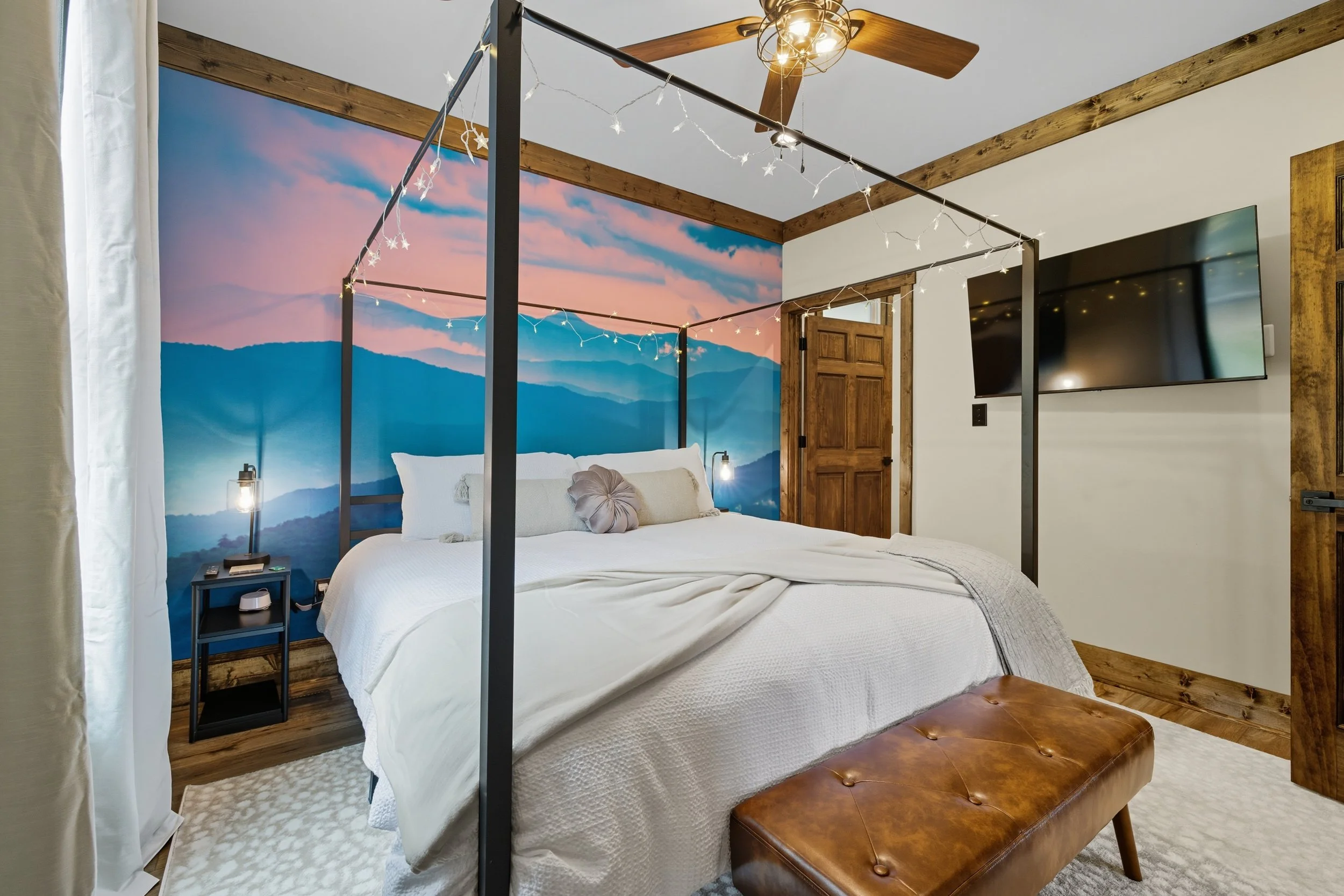 A bedroom with a large bed, string lights, a ceiling fan, a wall mural of mountains and sky, wooden accents, a flat-screen TV, and a small black bedside table with a lamp and books.