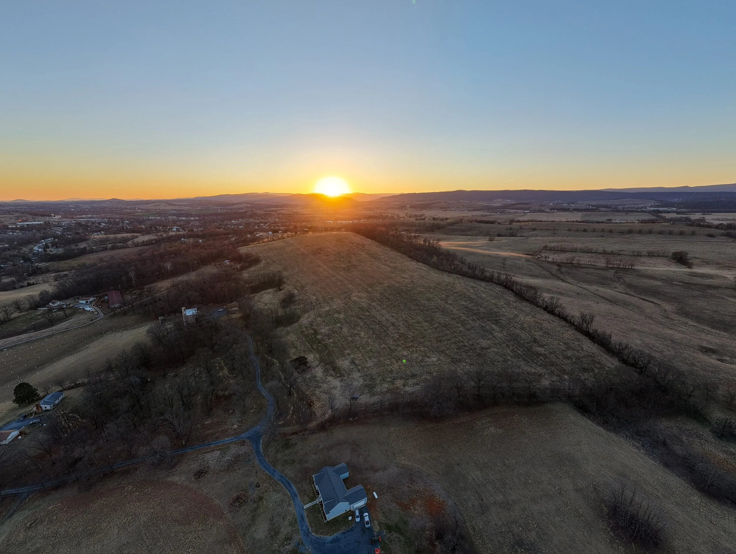 Aerial view of a rural landscape at sunset, showing open fields, scattered trees, a few houses, and winding roads under a clear sky.