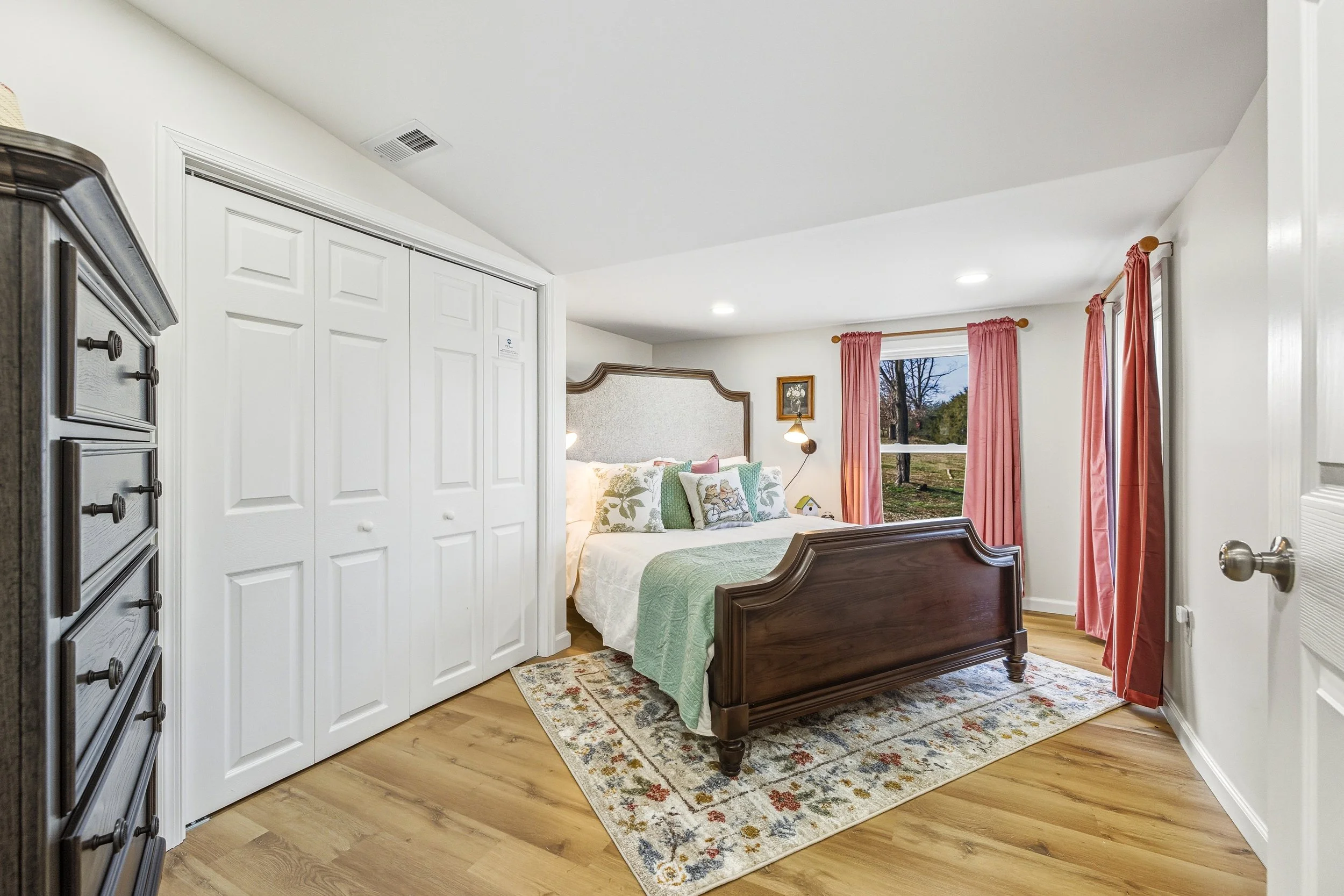 A cozy bedroom featuring a wooden bed with a green quilt and decorative pillows, pink curtains, a window showing a scenic outdoor view, a patterned rug, and a dresser on the left side.