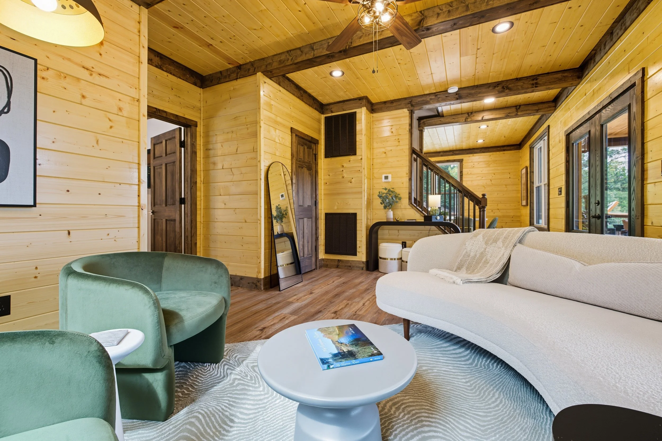 Interior of a cozy living room with wood-paneled walls and ceiling, featuring a white sofa, green armchair, and a round white coffee table with a magazine on it. Large windows show a view of greenery outside.