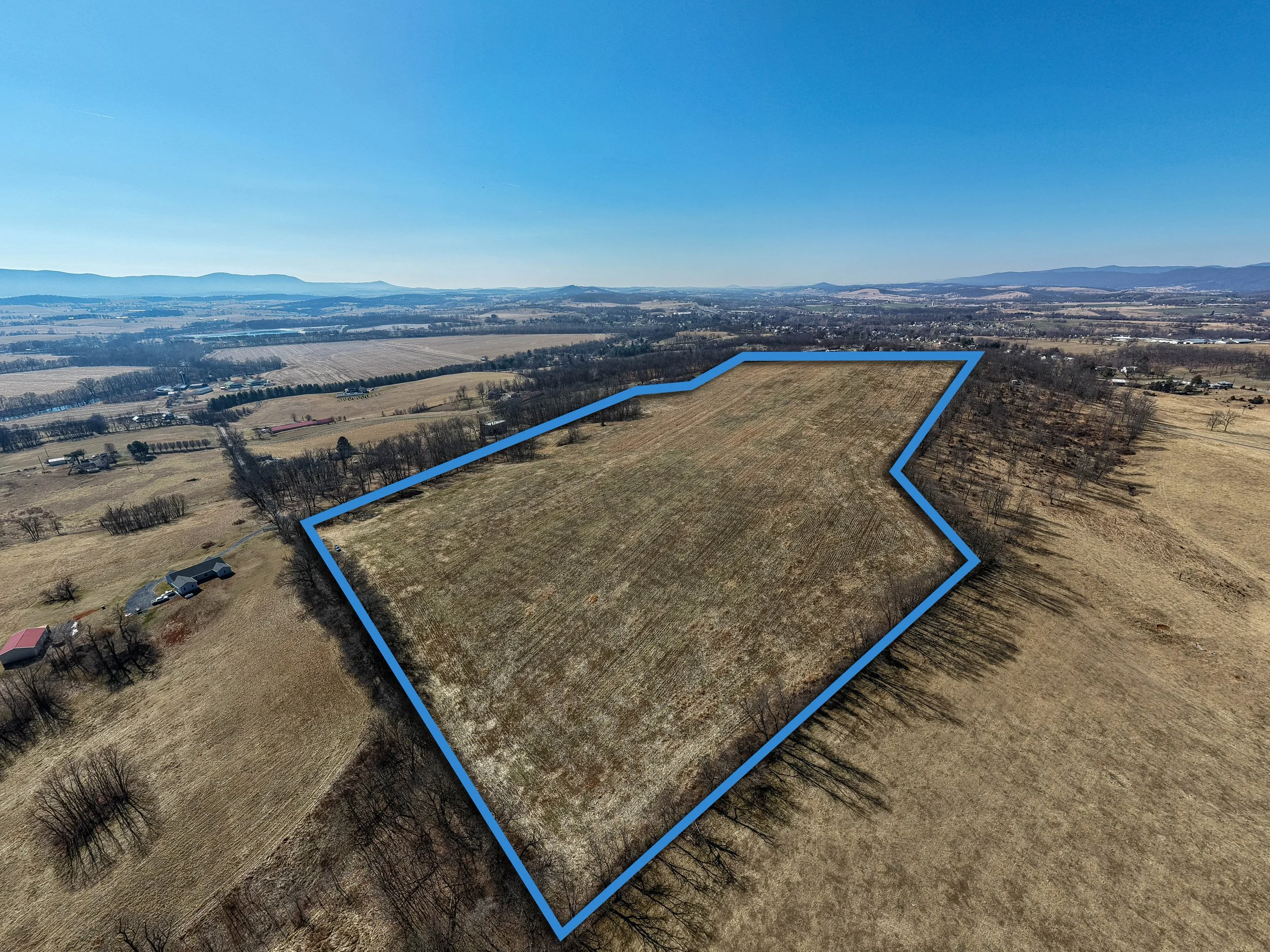 Aerial view of a rural landscape with a plot of land outlined in blue, fields, trees, and mountains in the background under a clear blue sky.