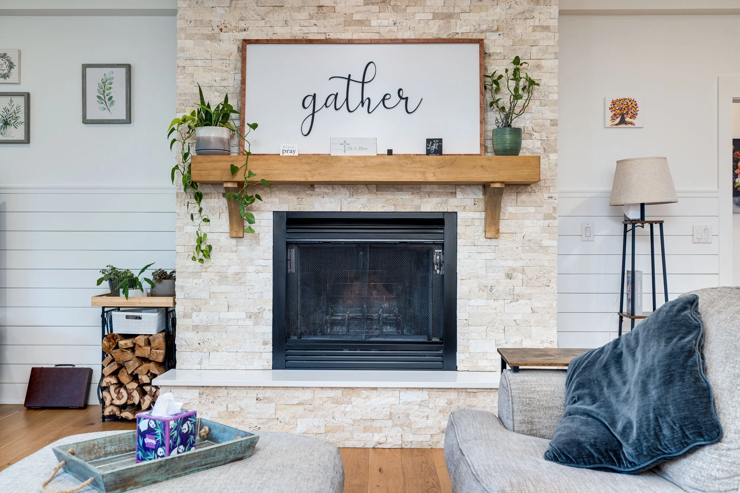 Living room fireplace with a wooden mantel, decorated with plants on both sides, a large framed sign reading 'gather' above, and wall art with trees and leaves on white walls. Furniture includes part of a gray armchair with a blue pillow and a coffee