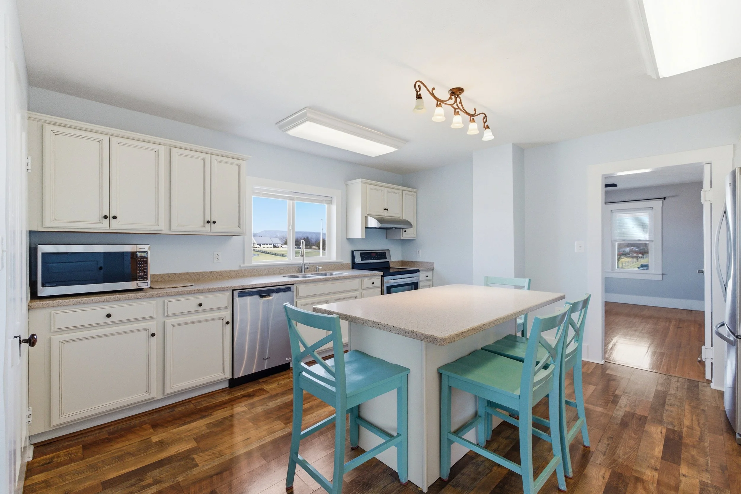 Bright kitchen with white cabinets, beige countertops, and a central island with turquoise chairs. Includes a window overlooking a green landscape, stainless steel appliances, and wooden flooring.