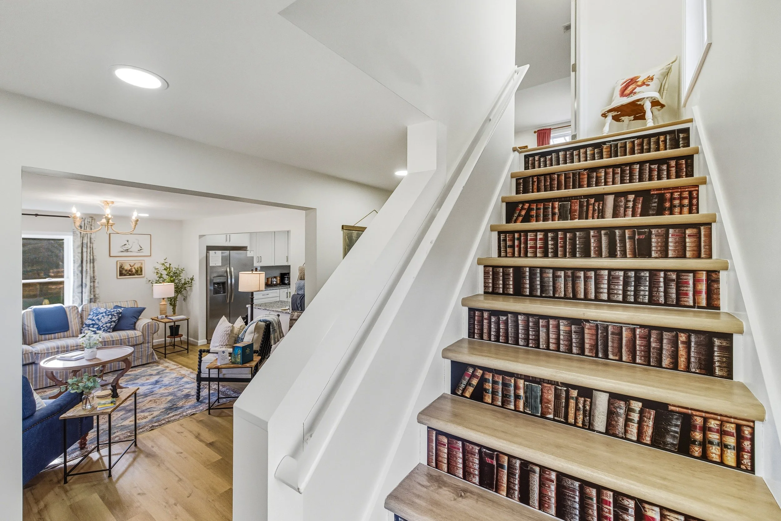 Living room with striped sofa, blue armchair, wooden coffee table, side tables, lamps, and artwork on the walls, visible from stairs with bookshelf designed as a book wall.