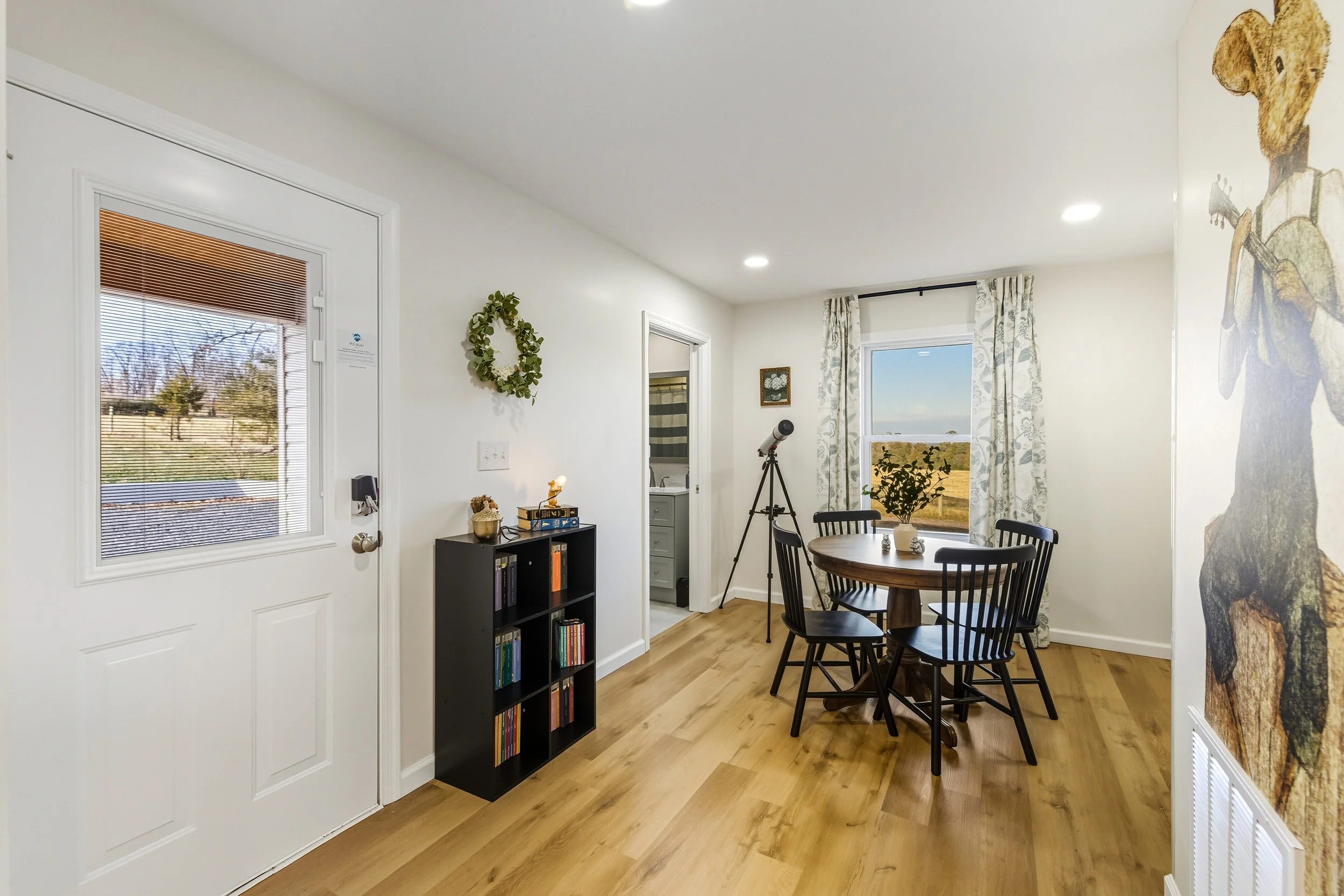A cozy dining area with a round wooden table surrounded by black chairs, a large window with floral curtains revealing an outdoor landscape, a telescope, a small bookshelf, and a wall mural of a bear playing a guitar.