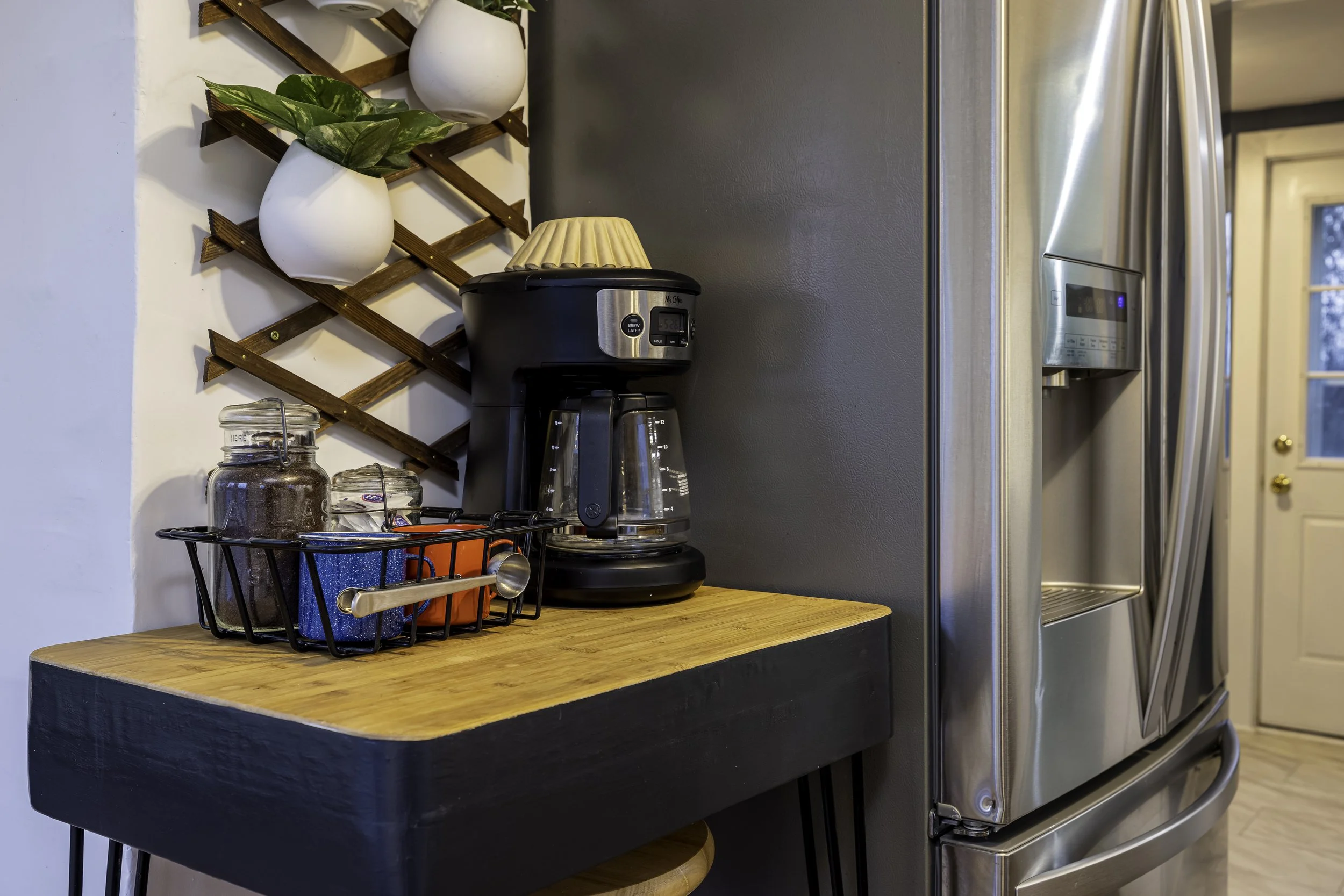 A kitchen counter with a black finish, featuring a coffee maker, a wire basket with jars and a orange container, and a wall-mounted wooden lattice with white planters holding green plants. To the right, a stainless steel refrigerator is visible.