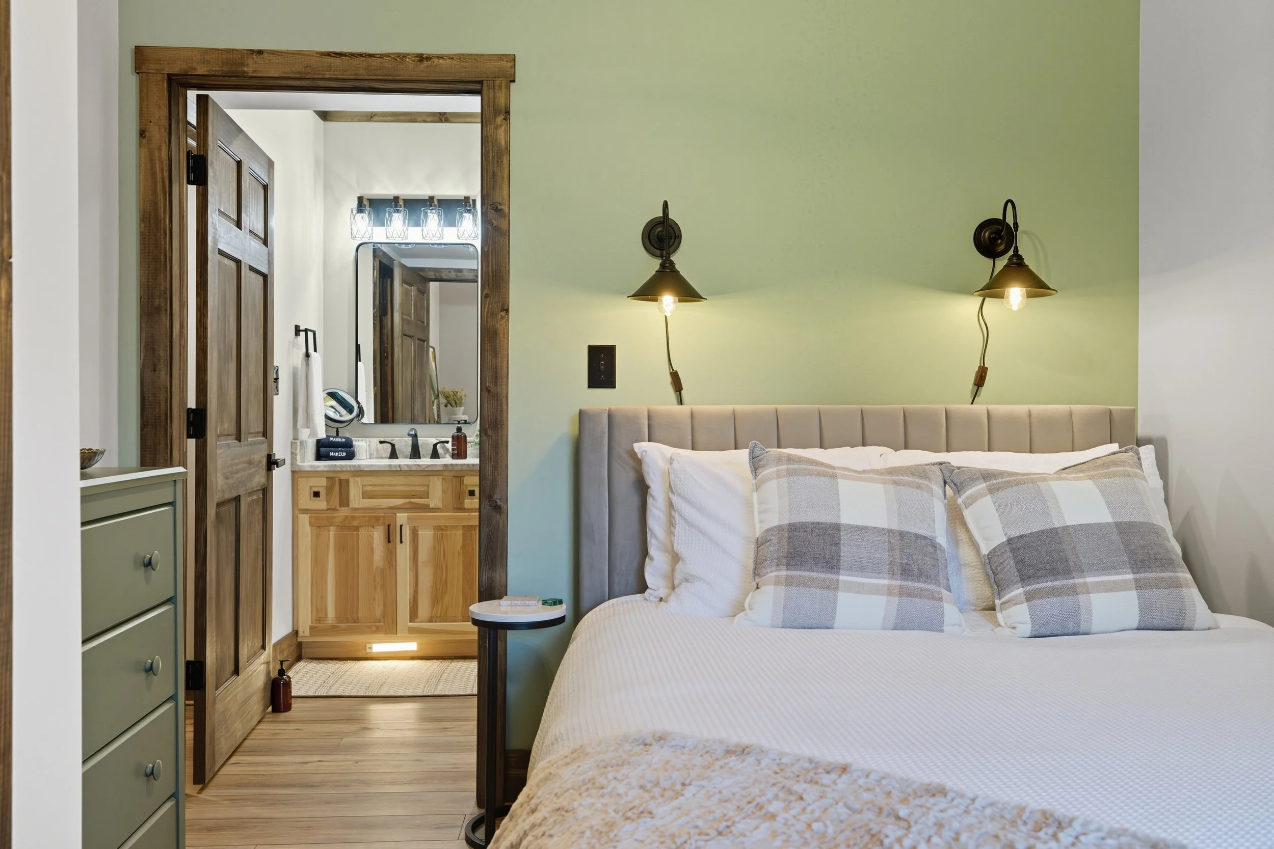 A bedroom with a bed that has a beige headboard and plaid pillows, two black wall-mounted lamps, and a nightstand with a small round table. An open doorway reveals a bathroom with a wooden vanity, a mirror, and a light fixture with several bulbs.