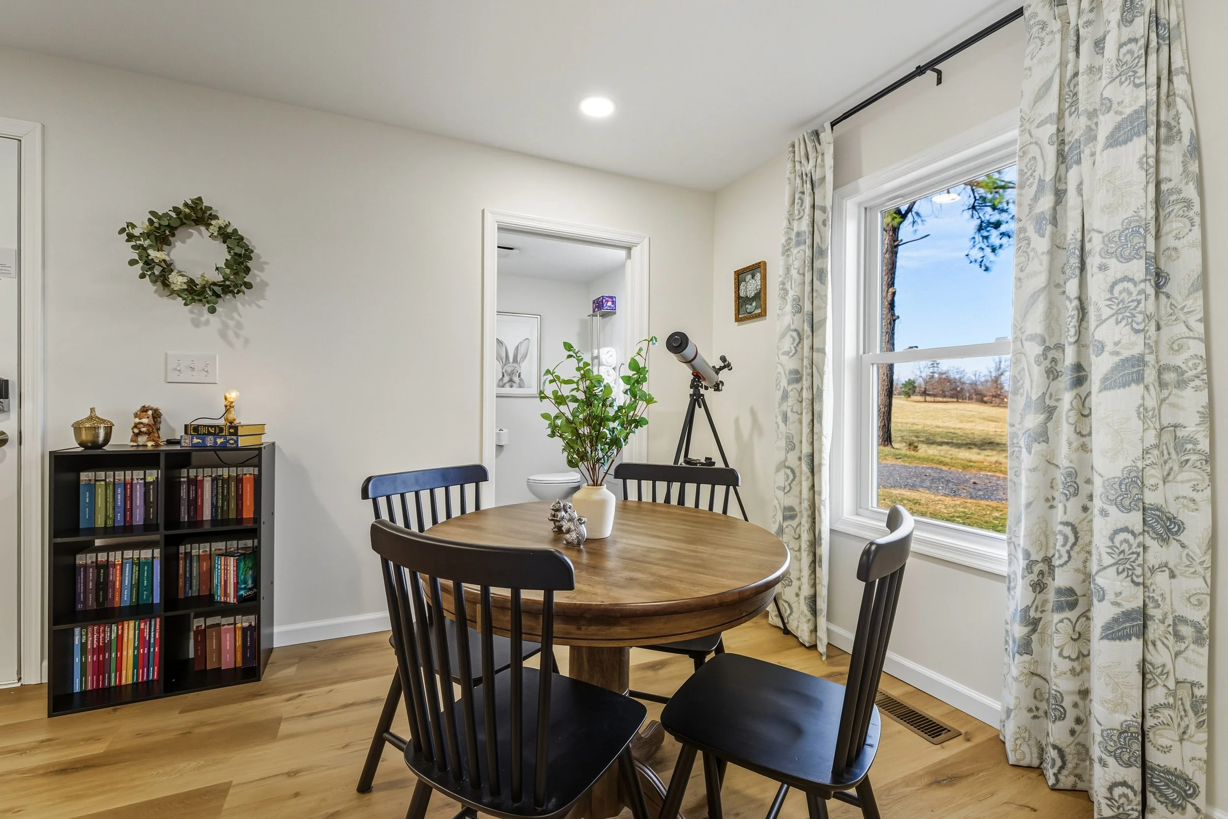 A dining room with a round wooden table and four black chairs, a window with floral curtains showing an outdoor scene with trees and grass, a bookshelf with colorful books, a small houseplant on the table, and a telescope nearby.