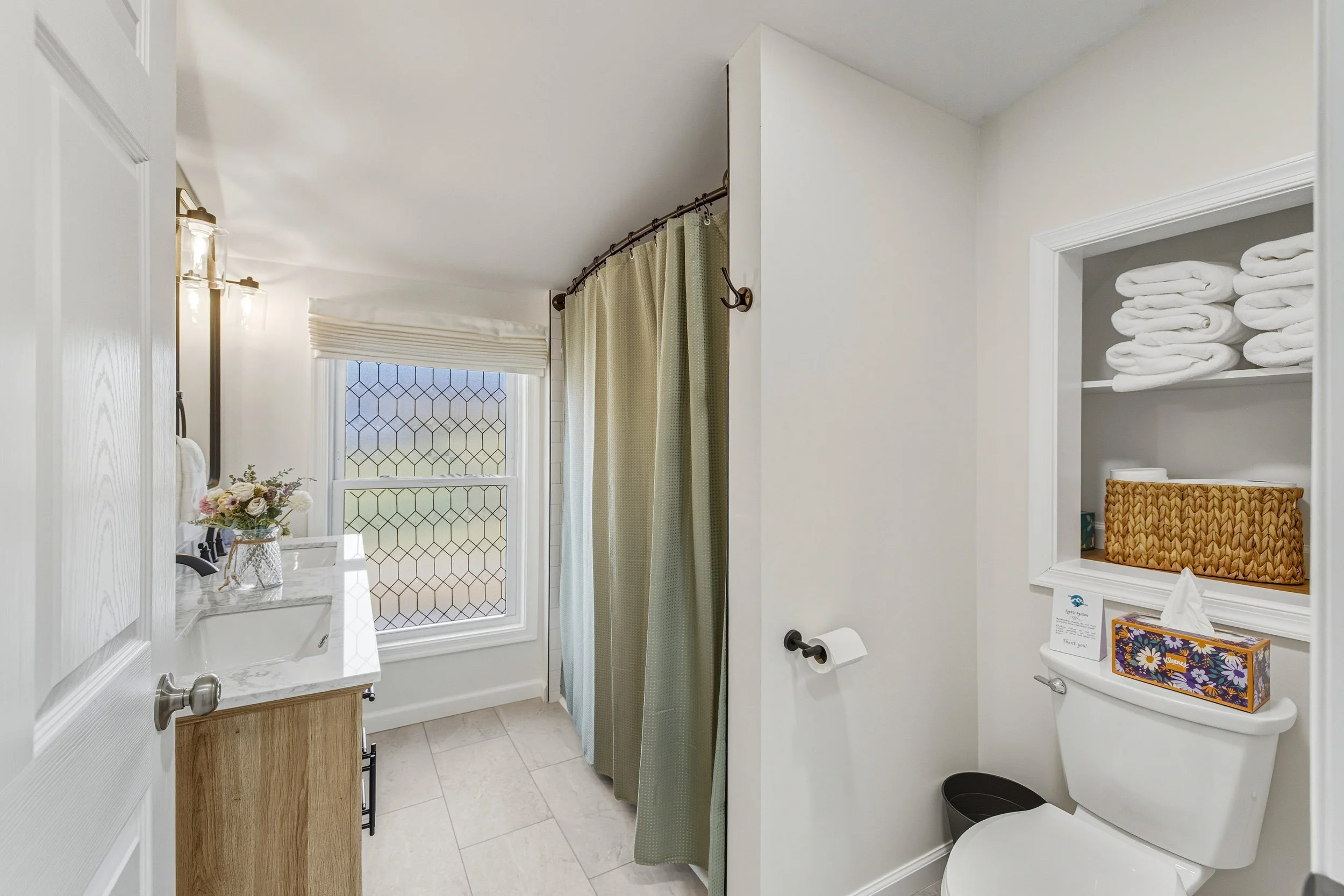 A bathroom with white walls, a window with diamond-patterned glass, a white marble sink, a bouquet of flowers, a shower with green curtains, a toilet with a tissue box on top, and shelves with white towels.