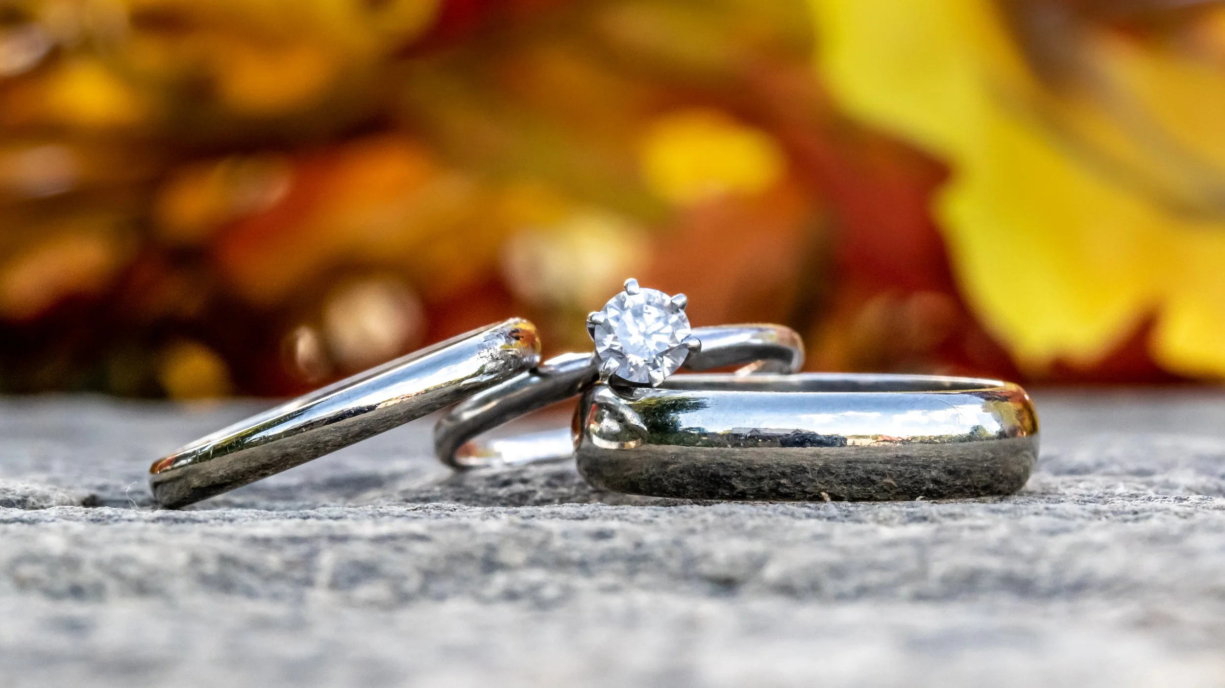 Close-up of two silver rings on a stone surface, one with a diamond, blurred floral background.