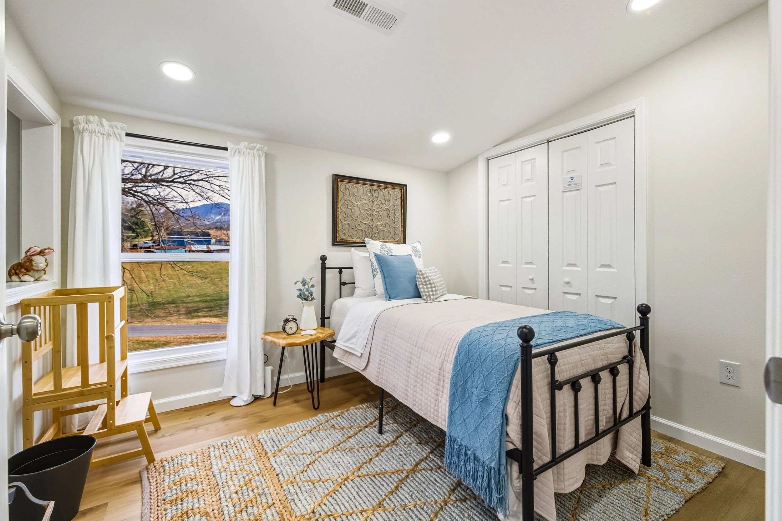 Comfortable bedroom with a metal bed, white and blue bedding, a side table with an alarm clock and lamp, a window with white curtains showing an outdoor view, a closet with white doors, decorative wall art, a rug, and a small wooden crib.