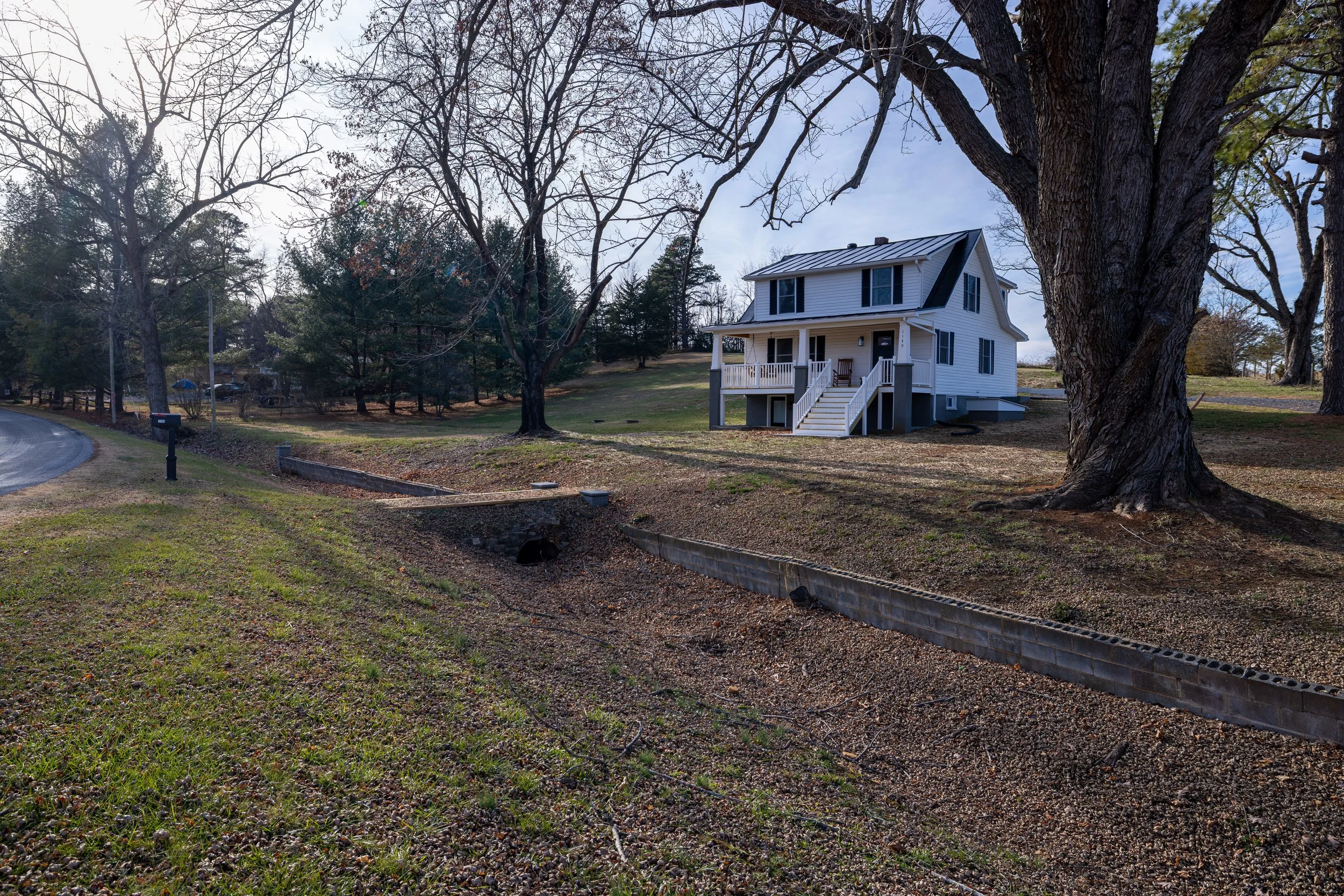 A suburban scene with a large tree in the foreground and a two-story white house with black shutters in the background. There are leafless trees and a cloudy sky.