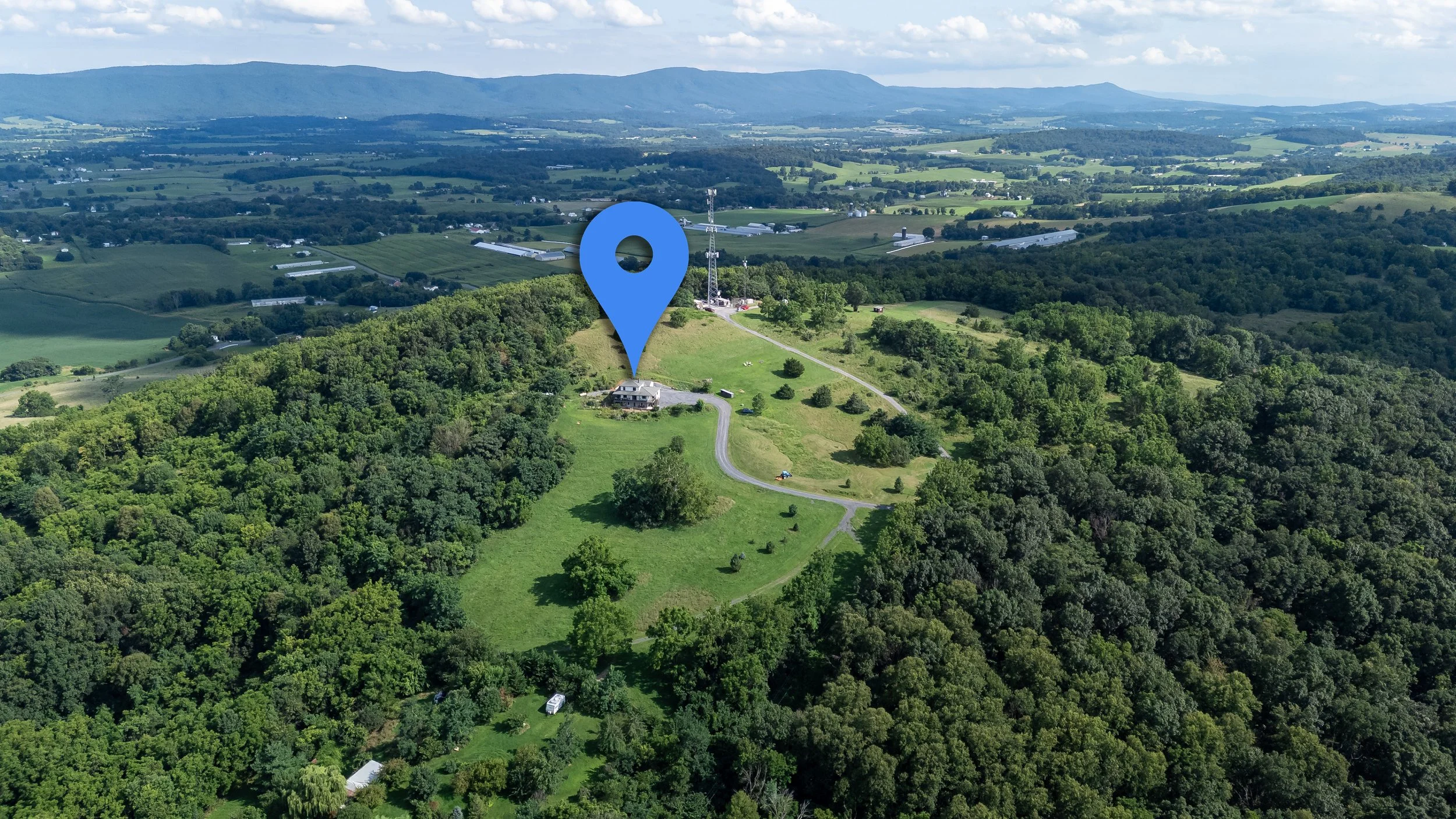 Aerial view of a rural landscape with rolling hills, green fields, dense forests, and a few scattered buildings. A large blue map pin marker is placed over a house near the top of a hill.