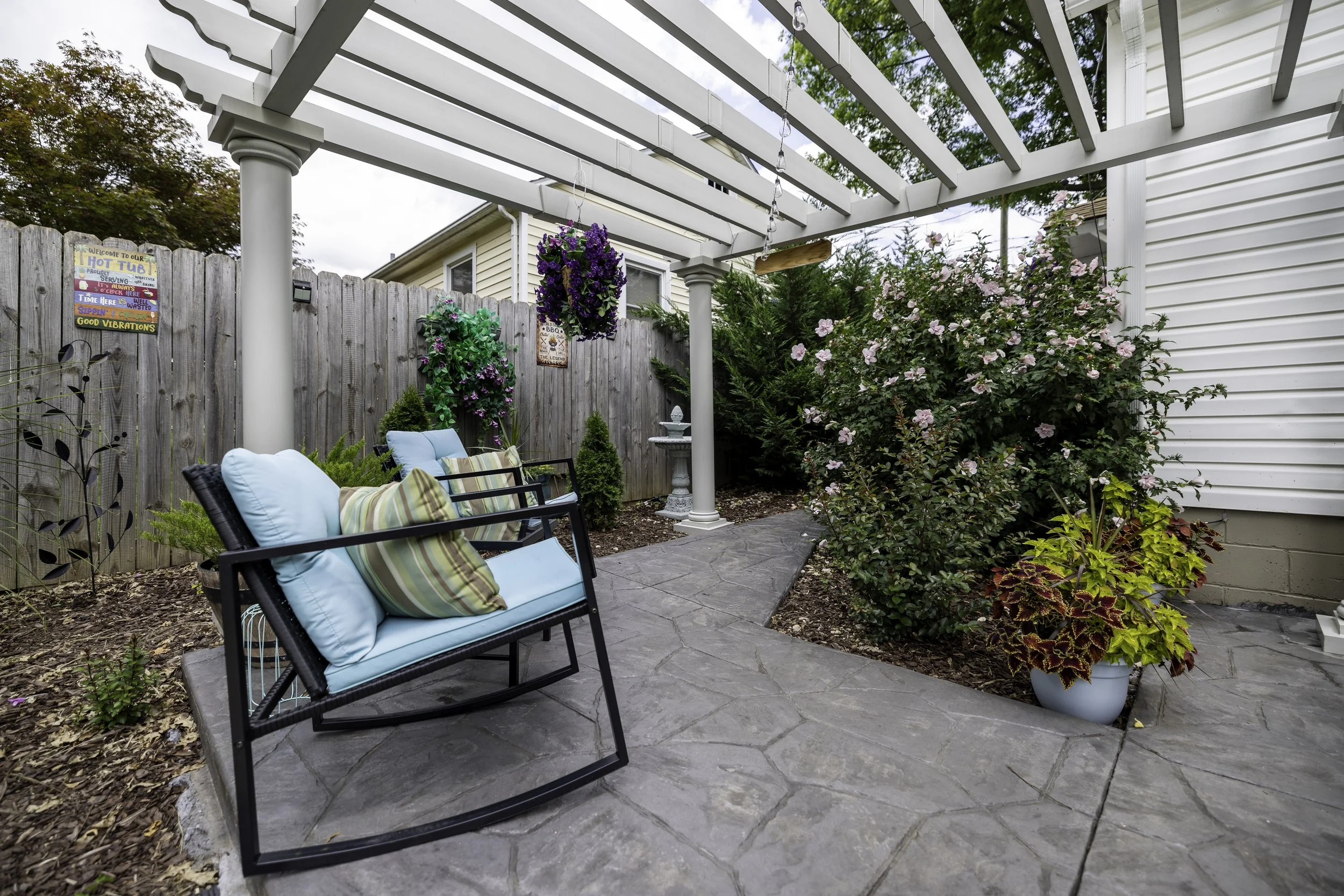 A backyard patio with two black chairs with light blue cushions and striped pillows, a gray stone pathway, a wooden fence, colorful flower decorations, a white pergola with hanging lights, various green plants, and a large flowering bush.