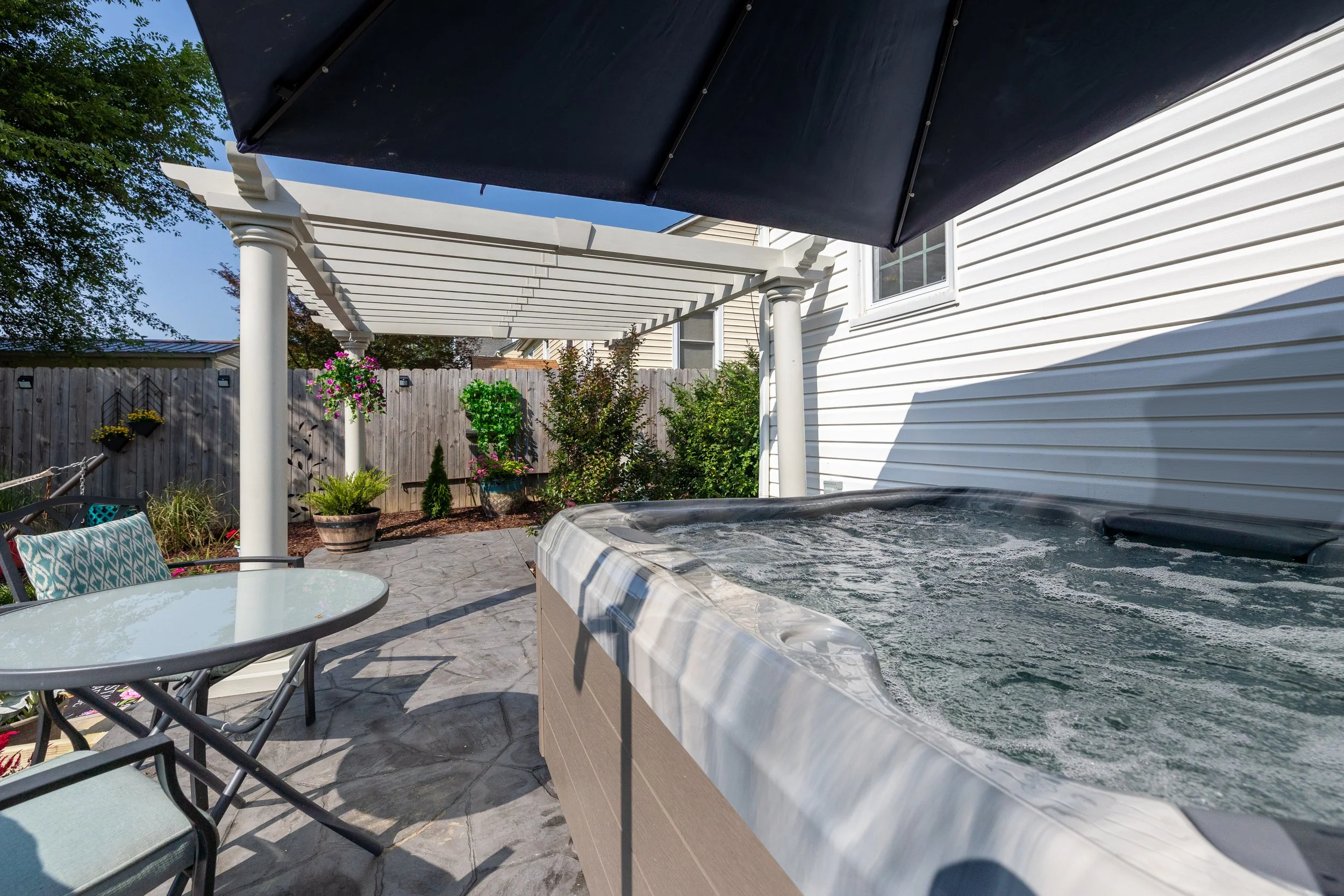 A backyard patio with a hot tub, patio furniture, potted plants, and a privacy fence with hanging flower baskets. There is a white house siding and a pergola with a dark umbrella overhead.