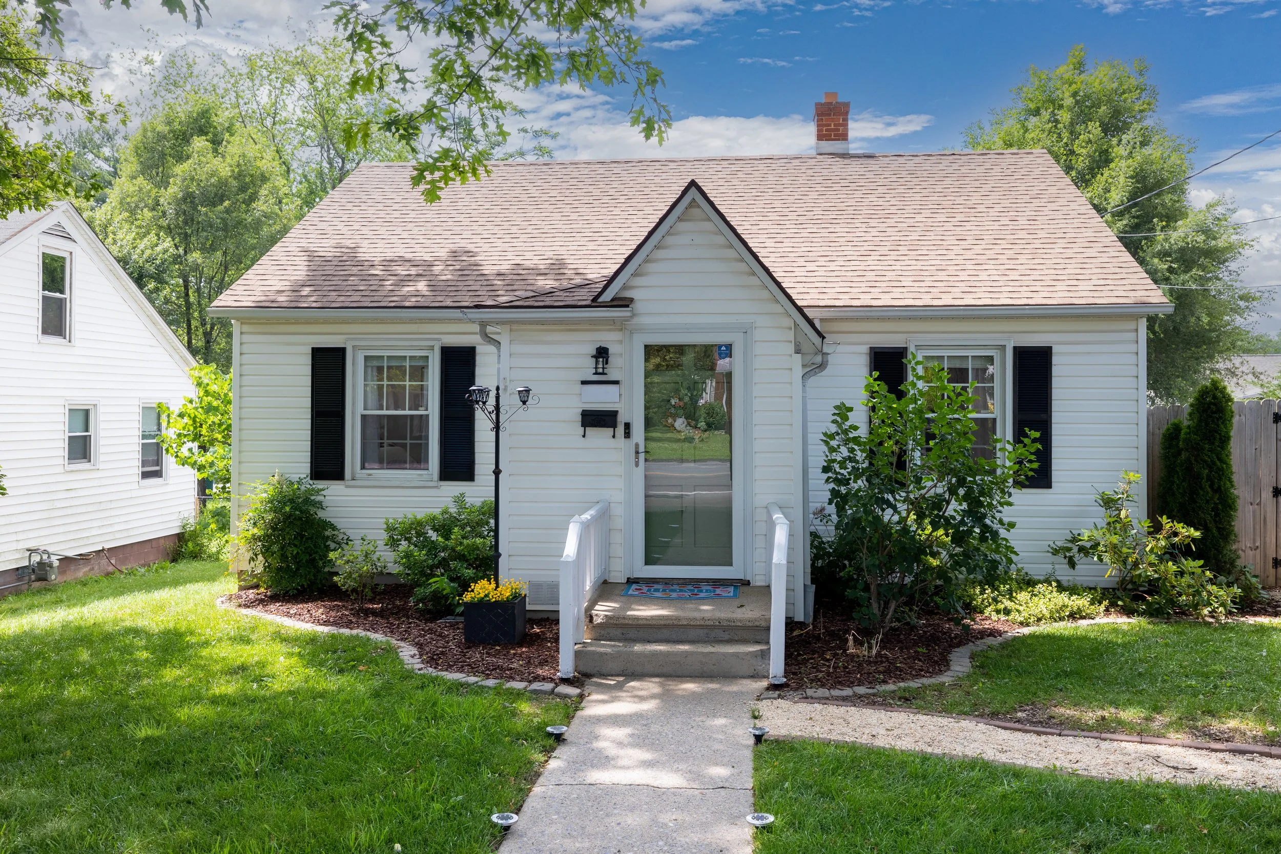 Front view of a small white house with black shutters, a porch with steps, and a glass door, surrounded by green plants and trees, under a partly cloudy sky.