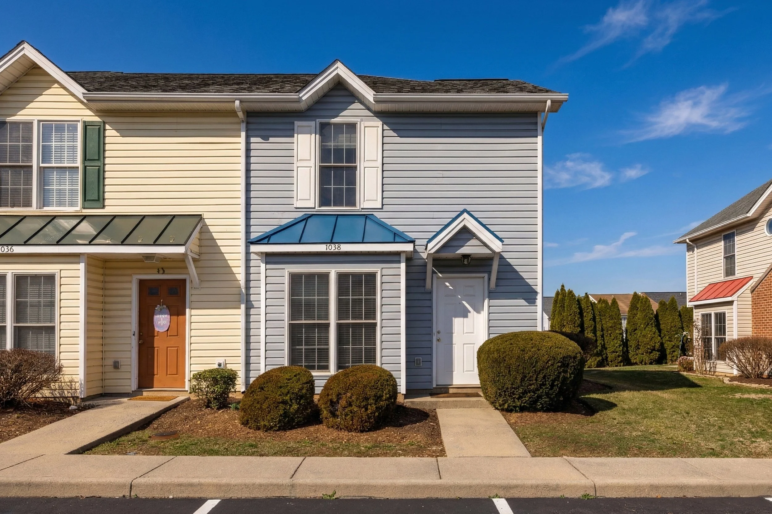 Front view of a row of townhouses with beige and blue siding, a small front yard with bushes, and a clear blue sky.