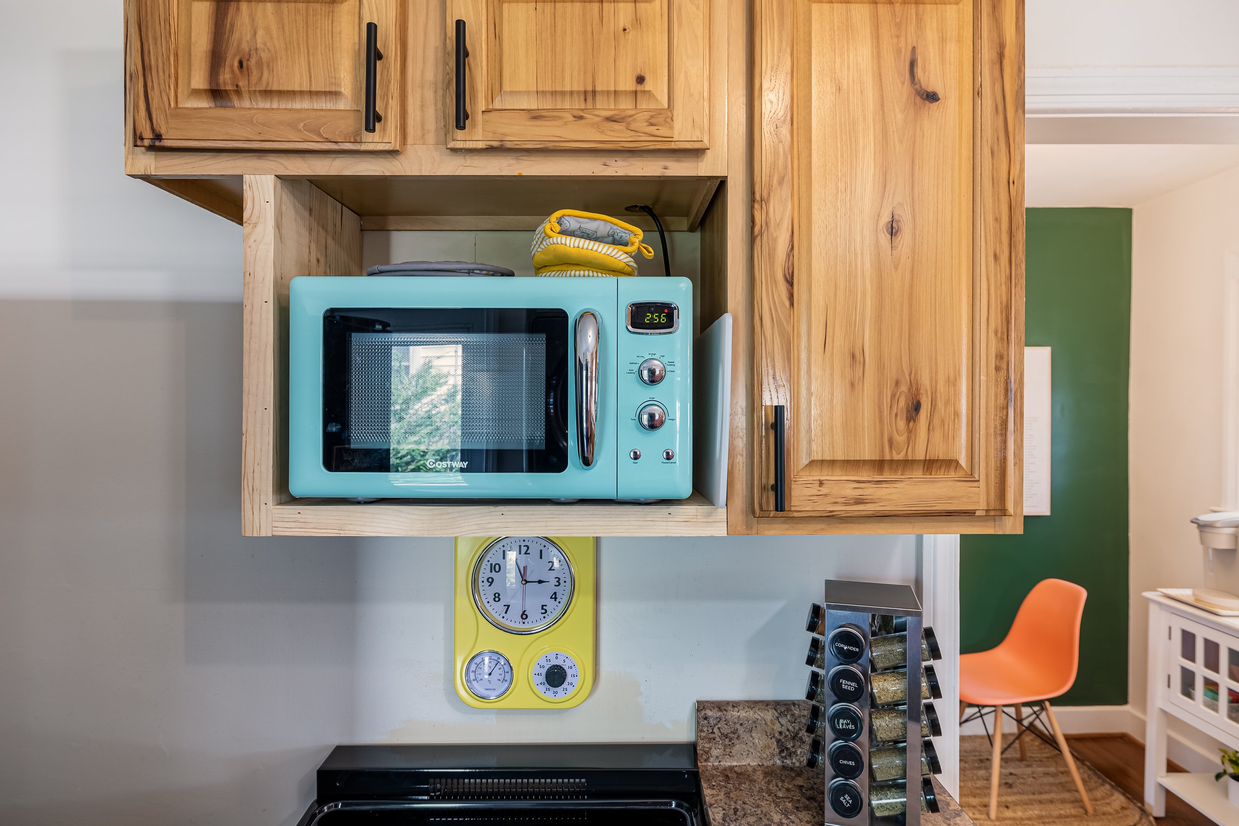 A kitchen countertop with a blue microwave oven, wooden cabinets, a yellow timer, a clock on the wall, and a spice rack with various spices.