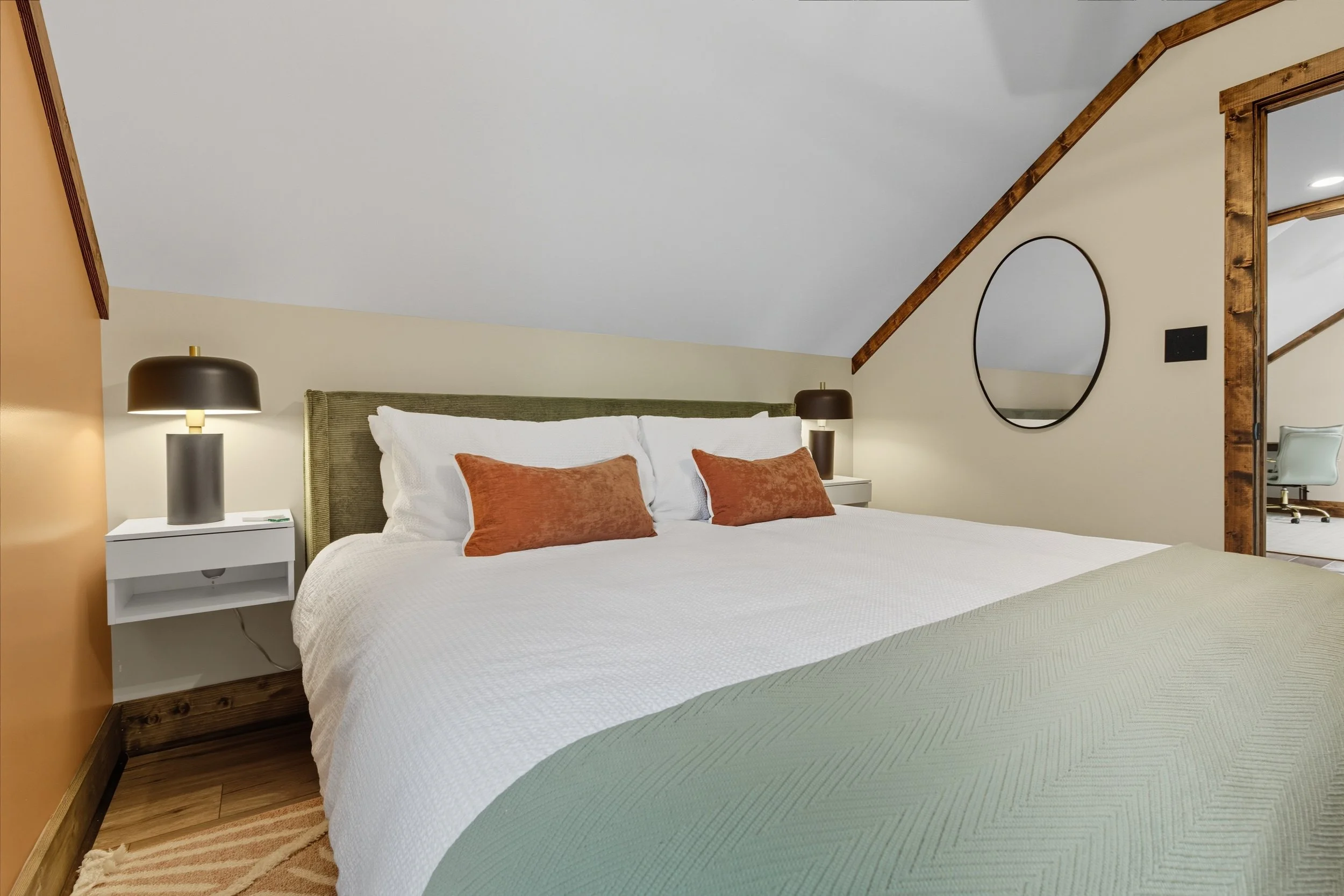 A cozy bedroom with a sloped ceiling and wooden trim, featuring a bed with white bedding and several rust-colored throw pillows, two black lamps on white nightstands, and a wall mirror with a black frame.
