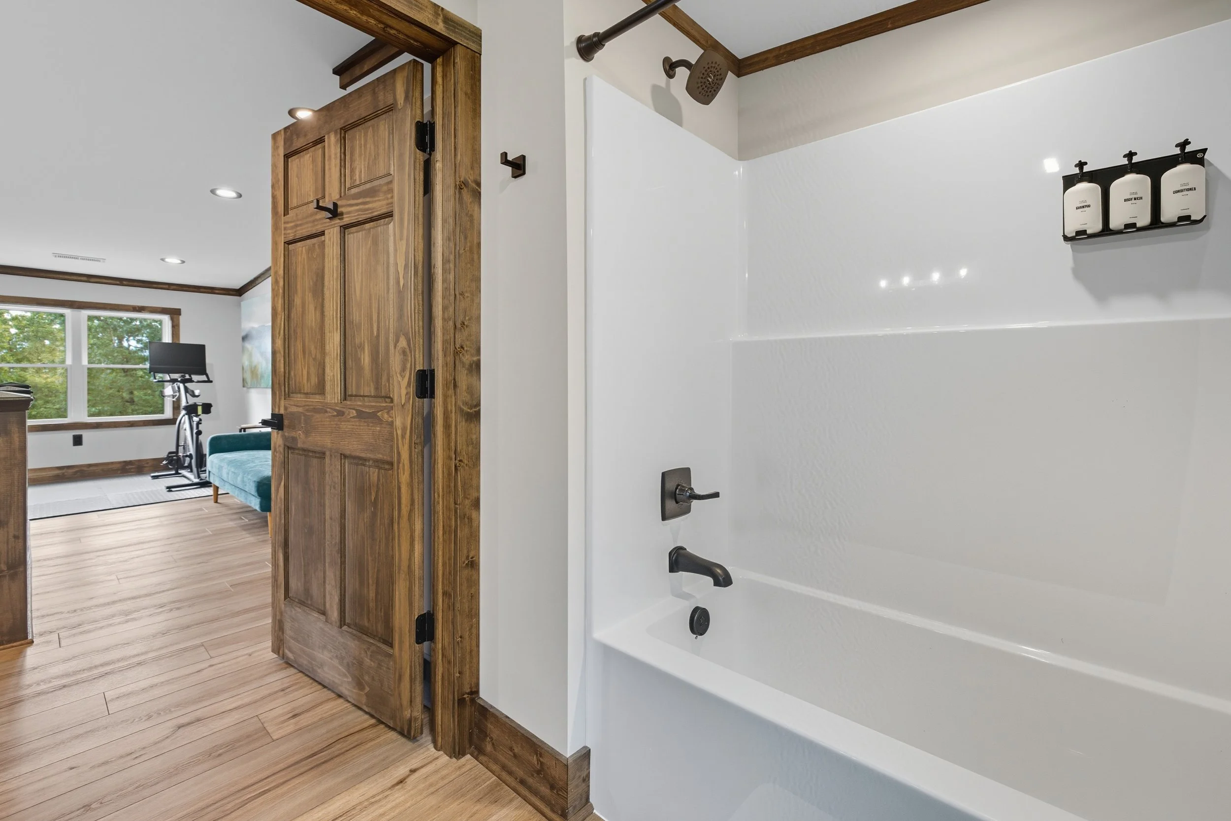A bathroom with a white bathtub, black fixtures, and a natural wood door leading to a room with a treadmill and windows with greenery outside.