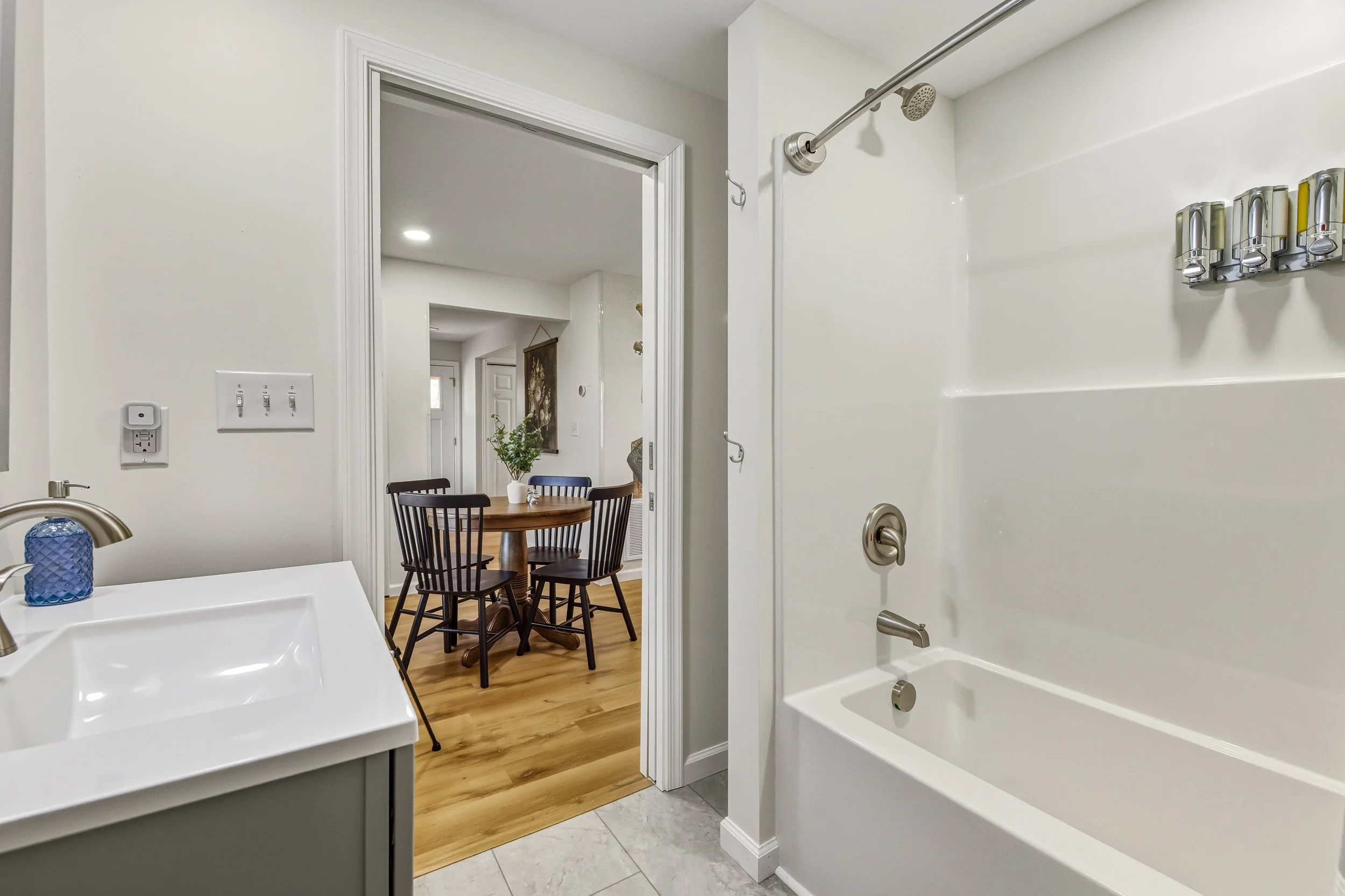 Bathroom with bathtub, shower, and sink. View into dining area with round table and chairs, visible through doorway.