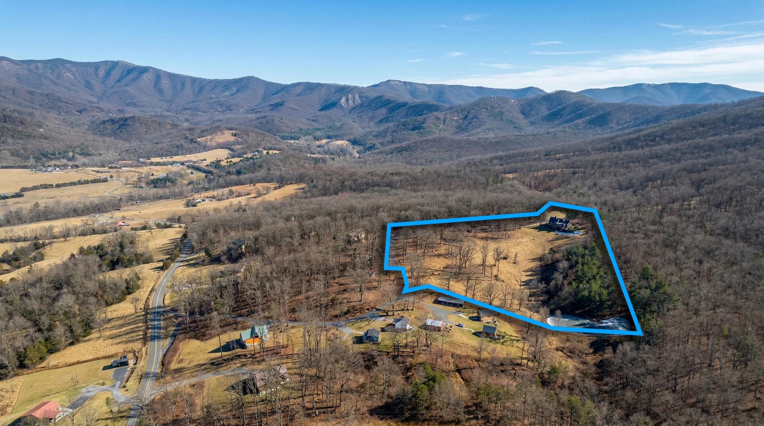 An aerial view of a rural landscape with rolling hills and mountains in the background, a property outlined with a blue line, and scattered houses and trees.
