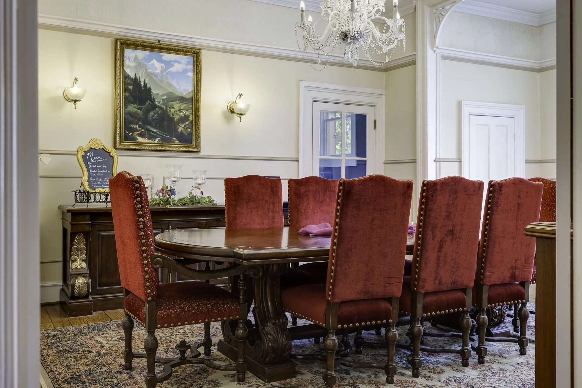 Elegant dining room with a large wooden table surrounded by red upholstered chairs with gold accents. A chandelier hangs from the ceiling, and there is a framed landscape painting on the wall. The room has cream-colored walls, white trim, and a window with outside greenery visible.