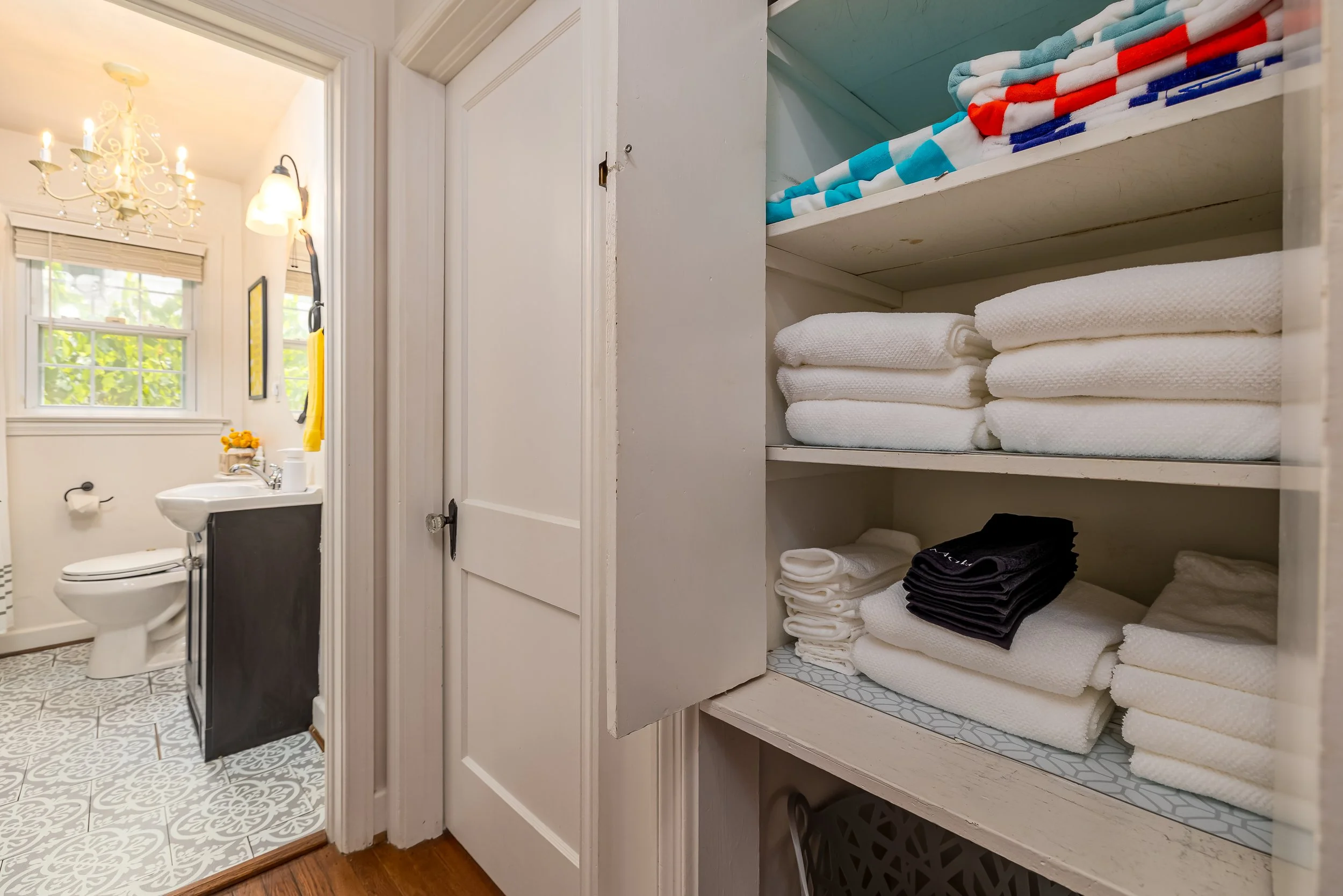 A view of a bathroom and linen closet. The bathroom has a white toilet, a small black cabinet, a sink, and a window with blinds. The linen closet has shelves with folded towels, including white, red, and blue striped towels, and darker towels at the 
