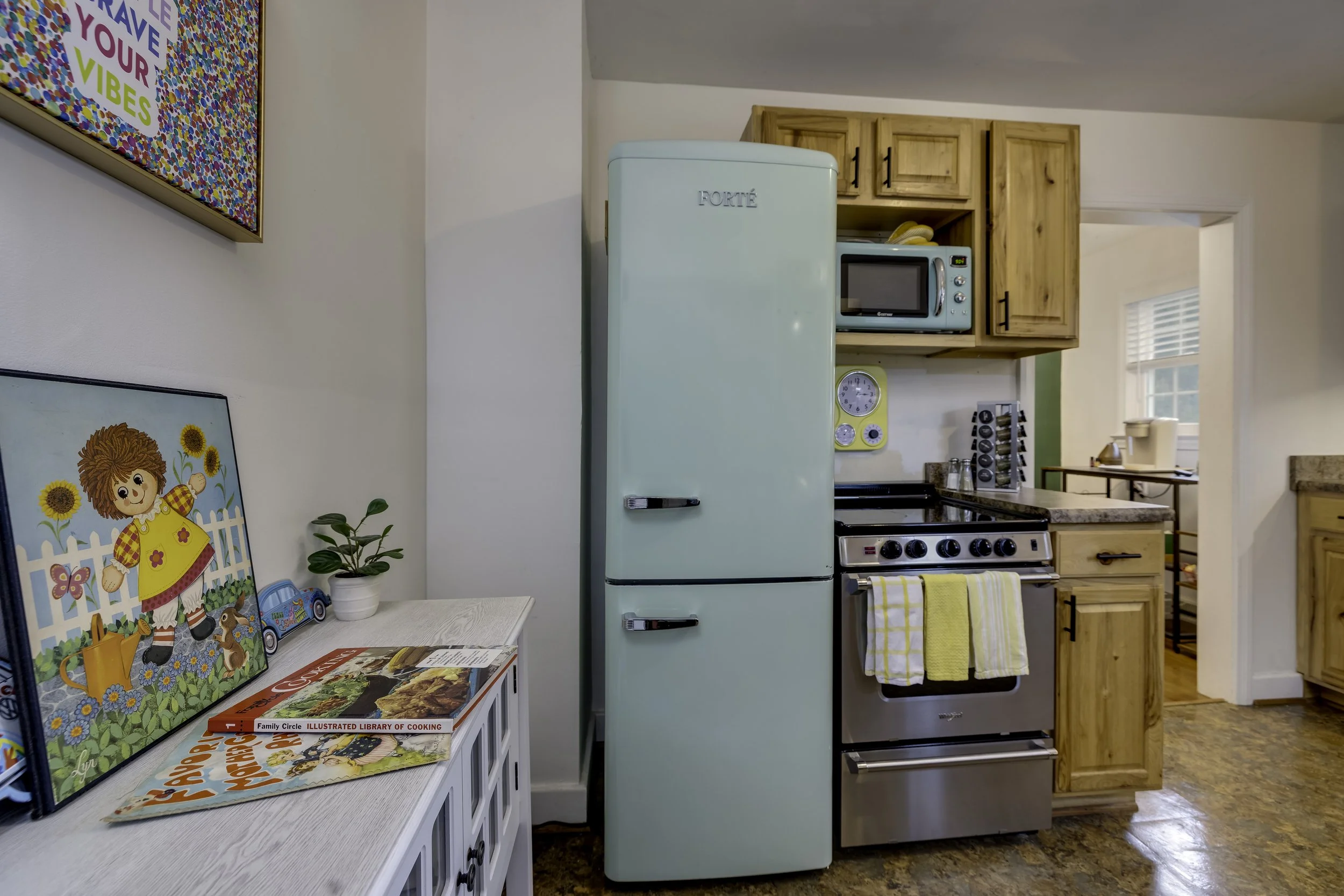A vintage mint green refrigerator in a kitchen with wooden cabinets, a stove, microwave, and colorful children's artwork on a white cabinet. There are books, plants, and a yellow towel in the scene.