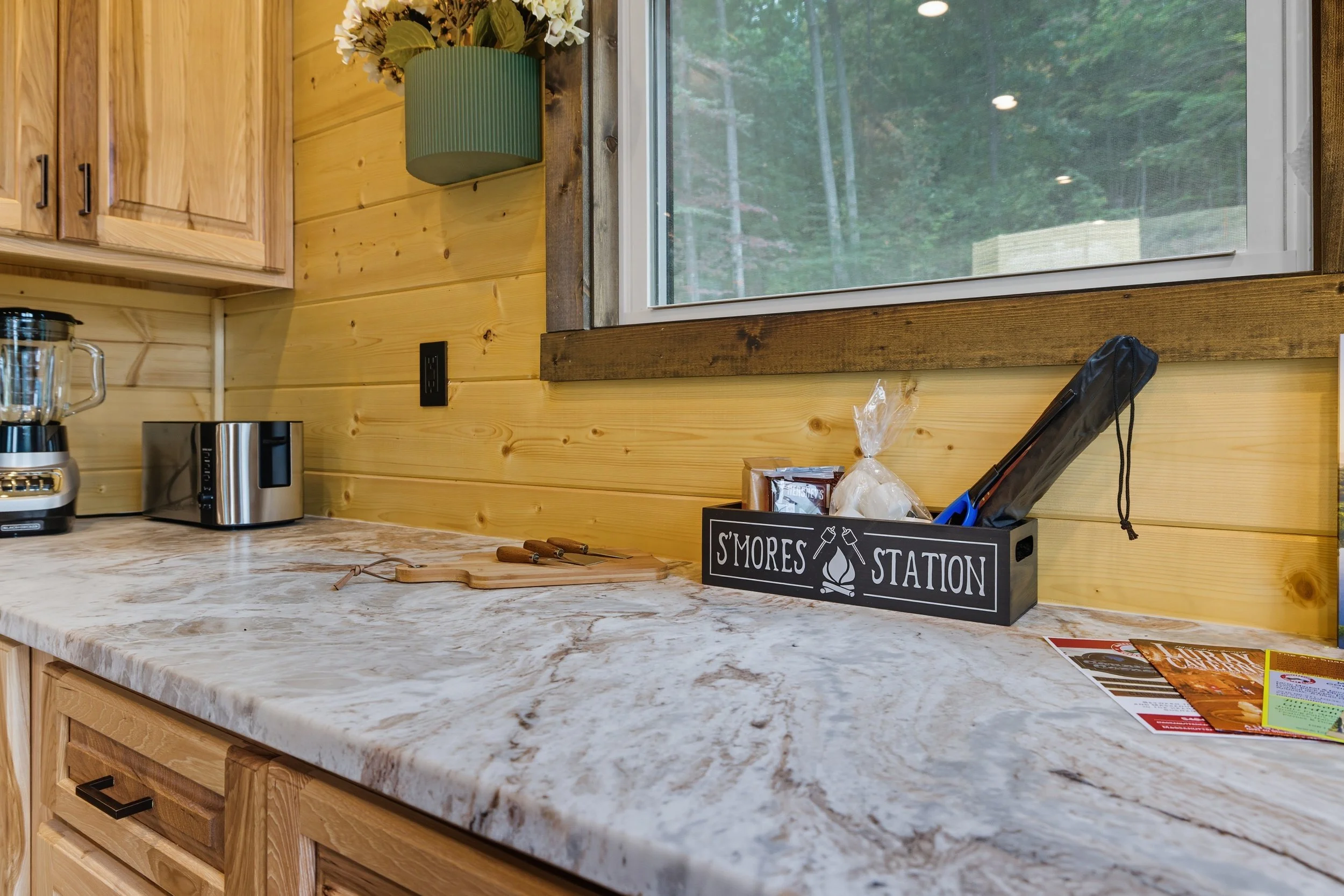 A kitchen countertop with a sign that says 'S'mores Station,' a cutting board, and a bag of marshmallows. The background includes wood-paneled walls, a window with a view of trees outside, and various kitchen appliances.
