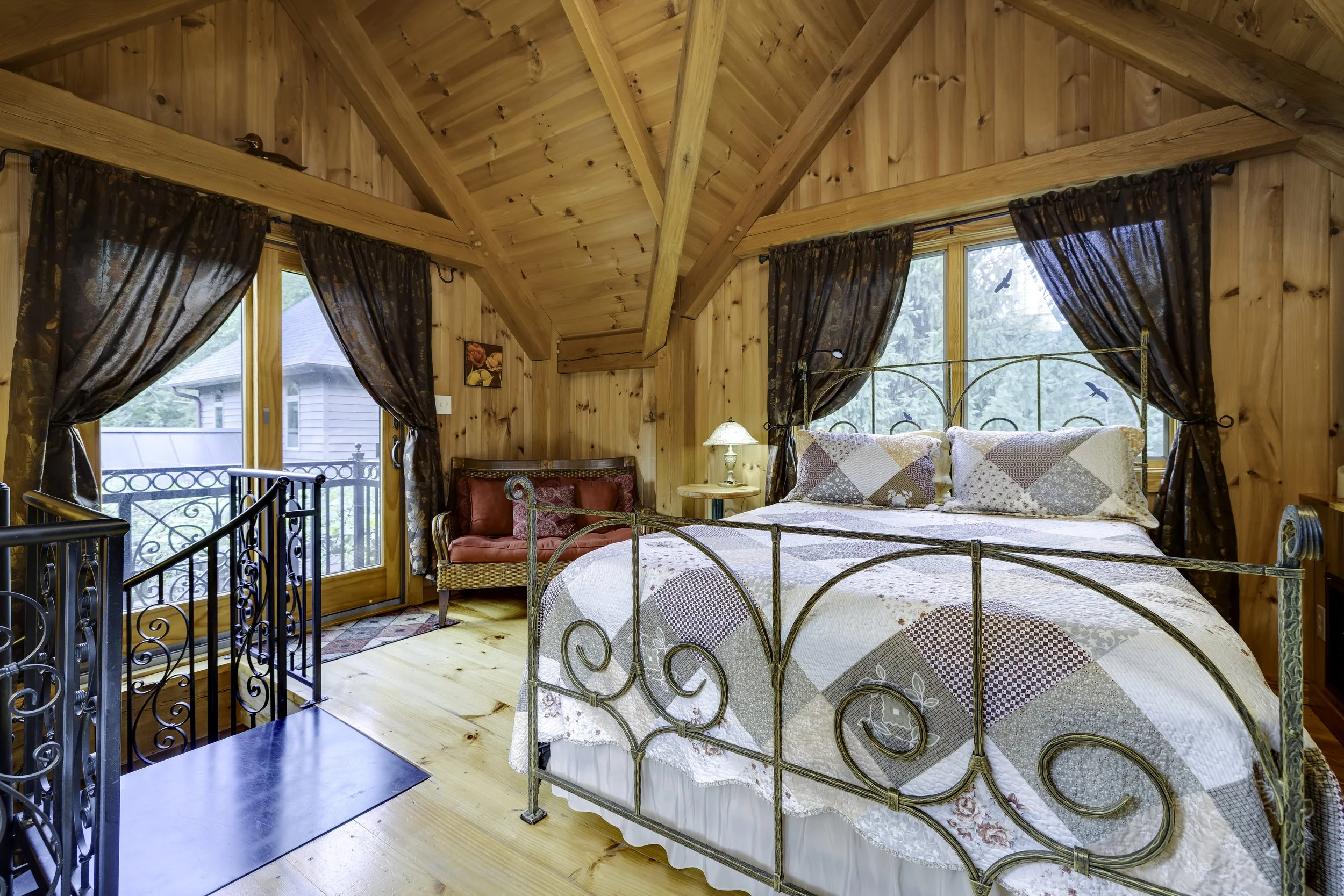 A cozy bedroom with wooden walls and ceiling, a metal bed with a quilt, dark patterned curtains on large windows, a small red armchair, a nightstand with a lamp, and a balcony with a view of trees.