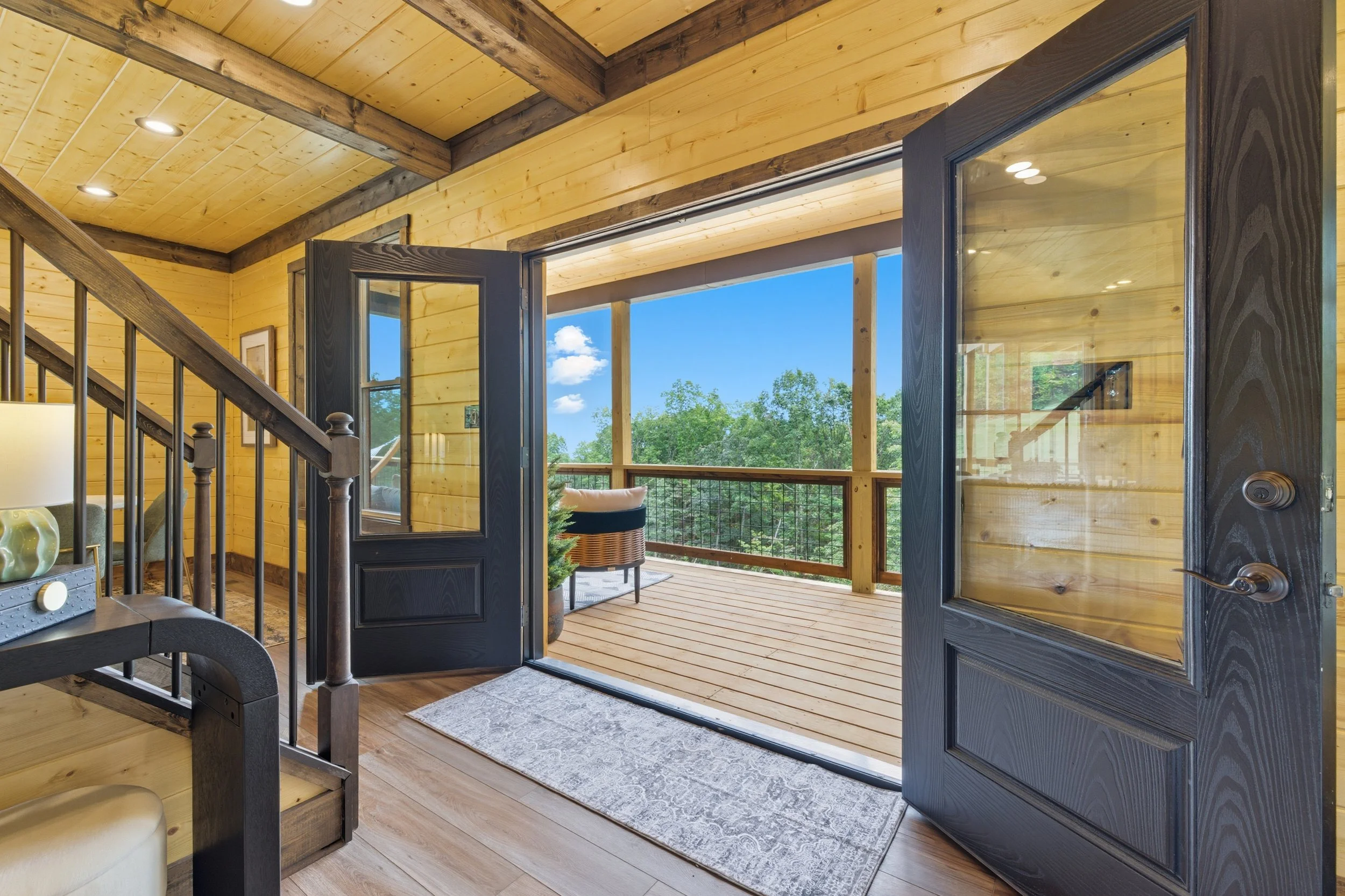 Open double doors leading onto a wooden porch with trees and blue sky in the background inside a rustic cabin with wood-paneled walls and ceiling.