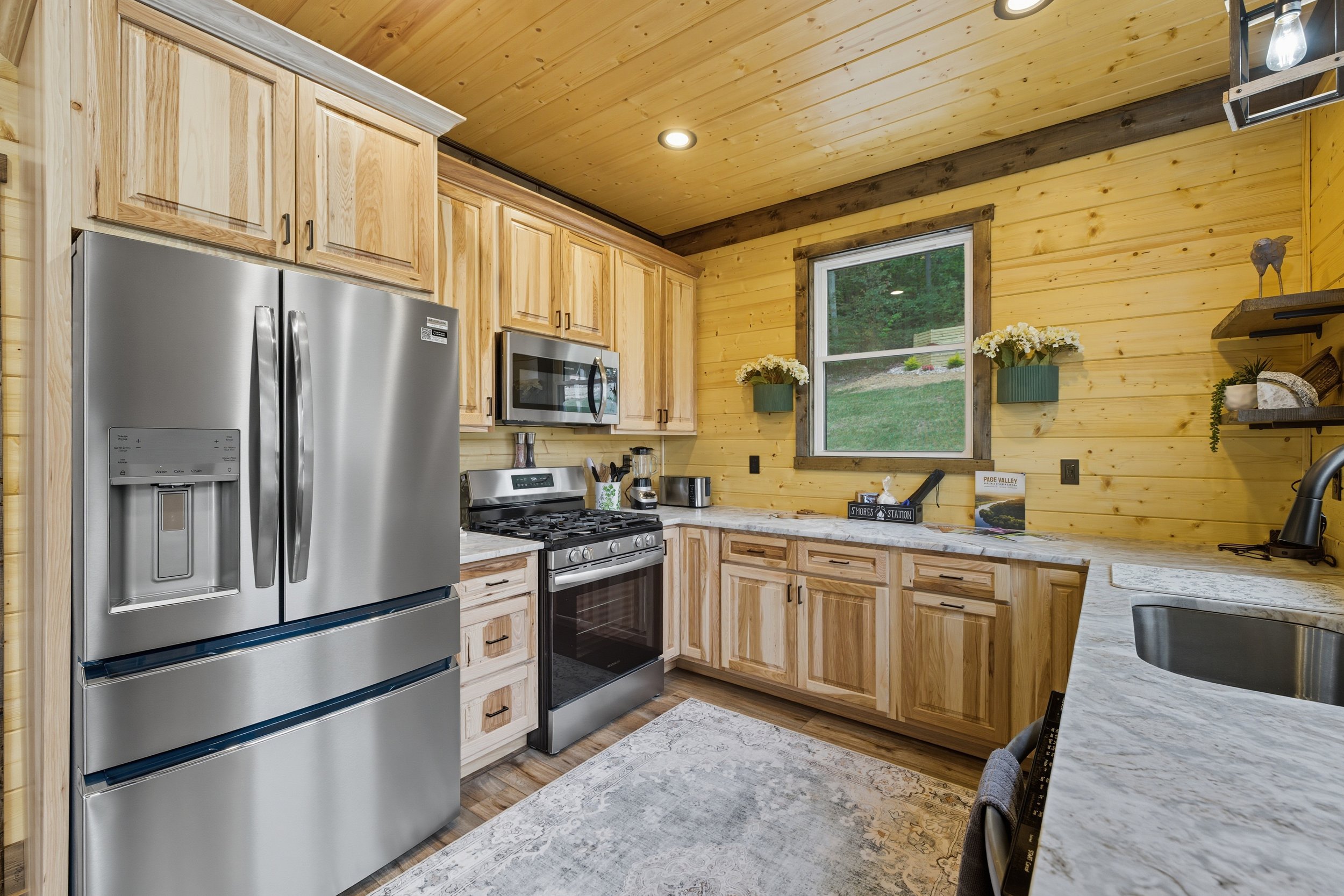 A kitchen with light wood cabinets, stainless steel refrigerator, microwave, dishwasher, and stove. It has a window and decorative wall shelves with plants and decor.