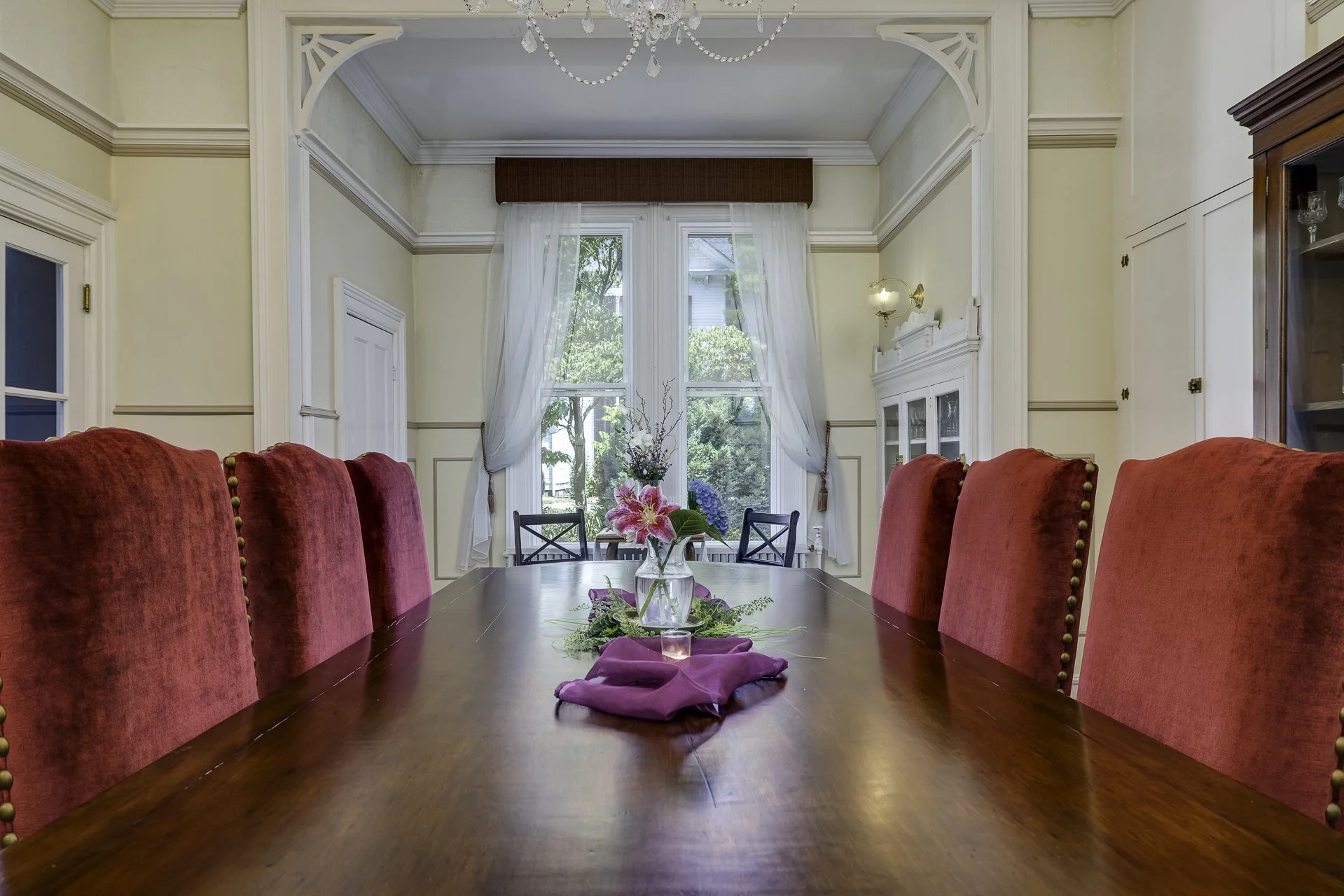 Dining room with a large wooden table surrounded by red upholstered chairs; a vase with pink lilies and greenery on the table, large windows with white curtains and greenery outside, decorative crown molding, a crystal chandelier, a white cabinet, and a tall wooden display cabinet.