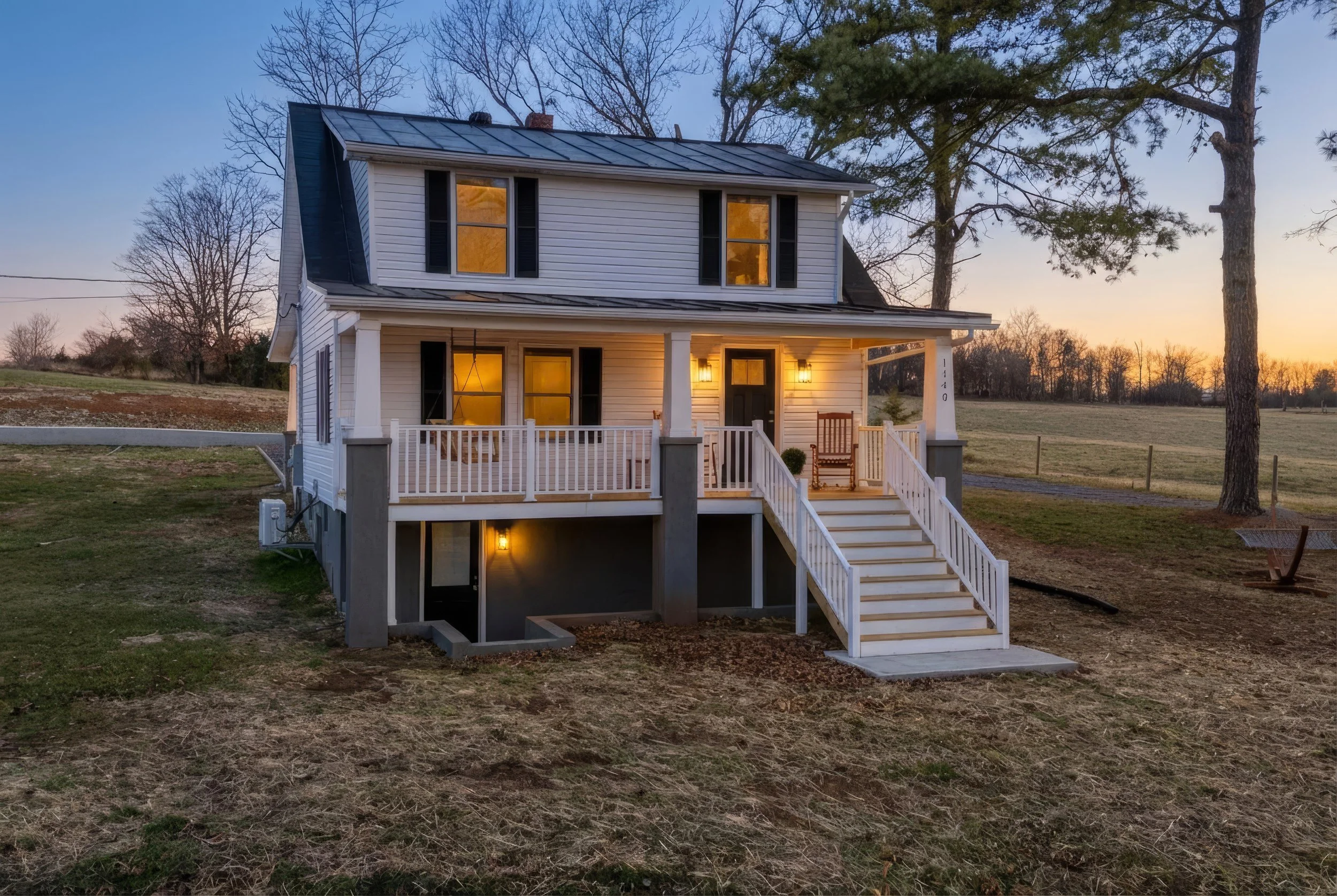 A two-story white house with black shutters and a porch, illuminated by warm lighting, viewed at dusk with a backdrop of trees and a sunset sky.