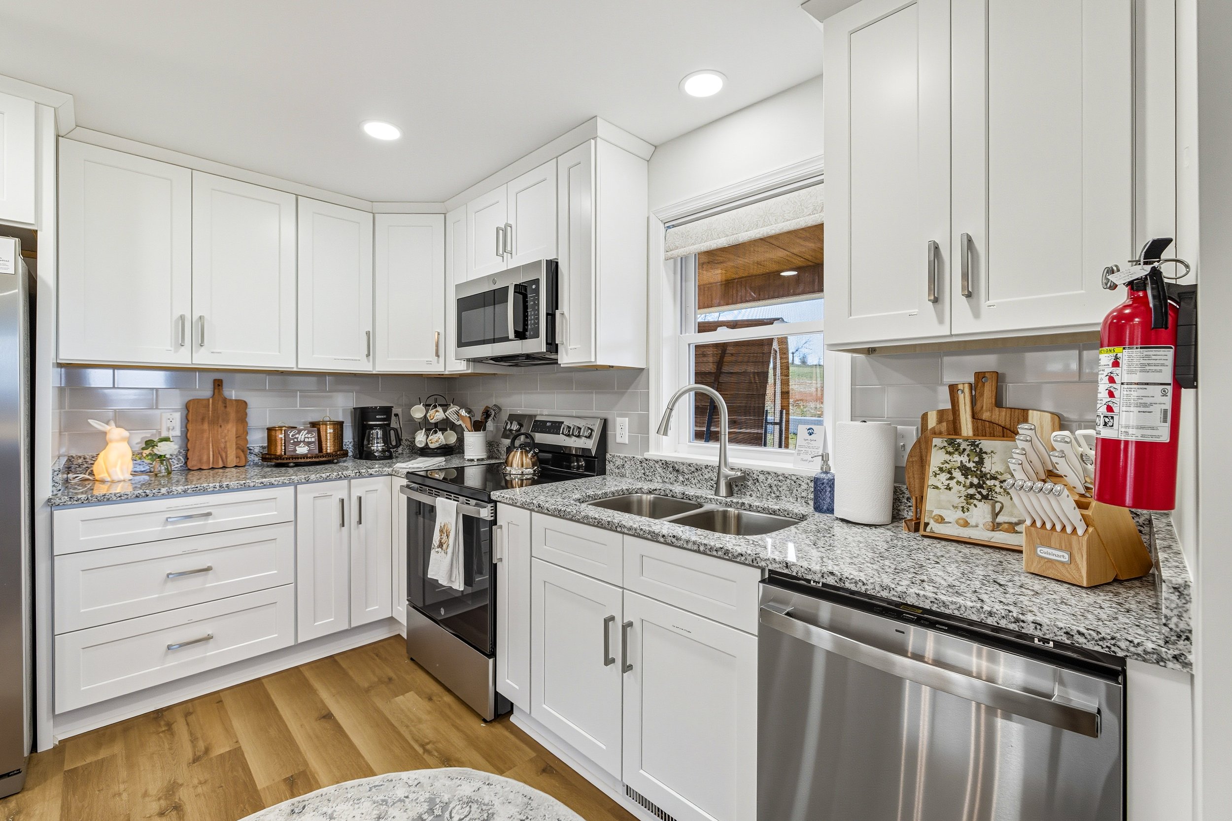 White kitchen with granite countertops, white cabinets, stainless steel stove and microwave, window above sink, fire extinguisher on the right, various kitchen utensils and decor on the counter.
