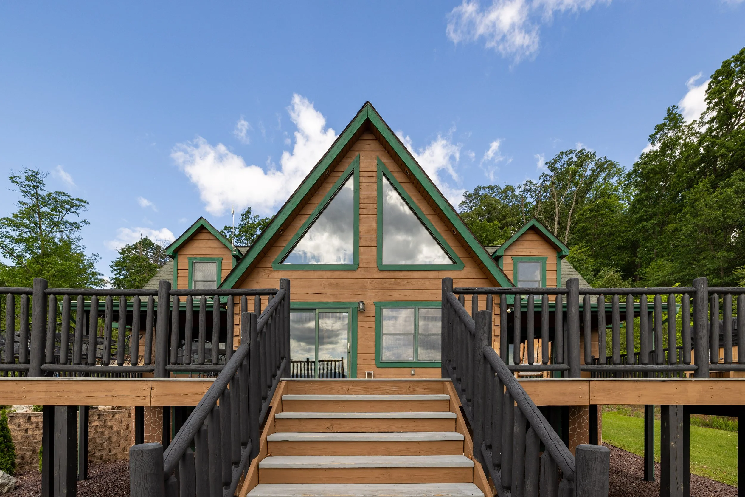 A wooden house with a prominent triangular front, green trim, and large window reflections, surrounded by a dark wooden fence and lush green trees under a partly cloudy sky.