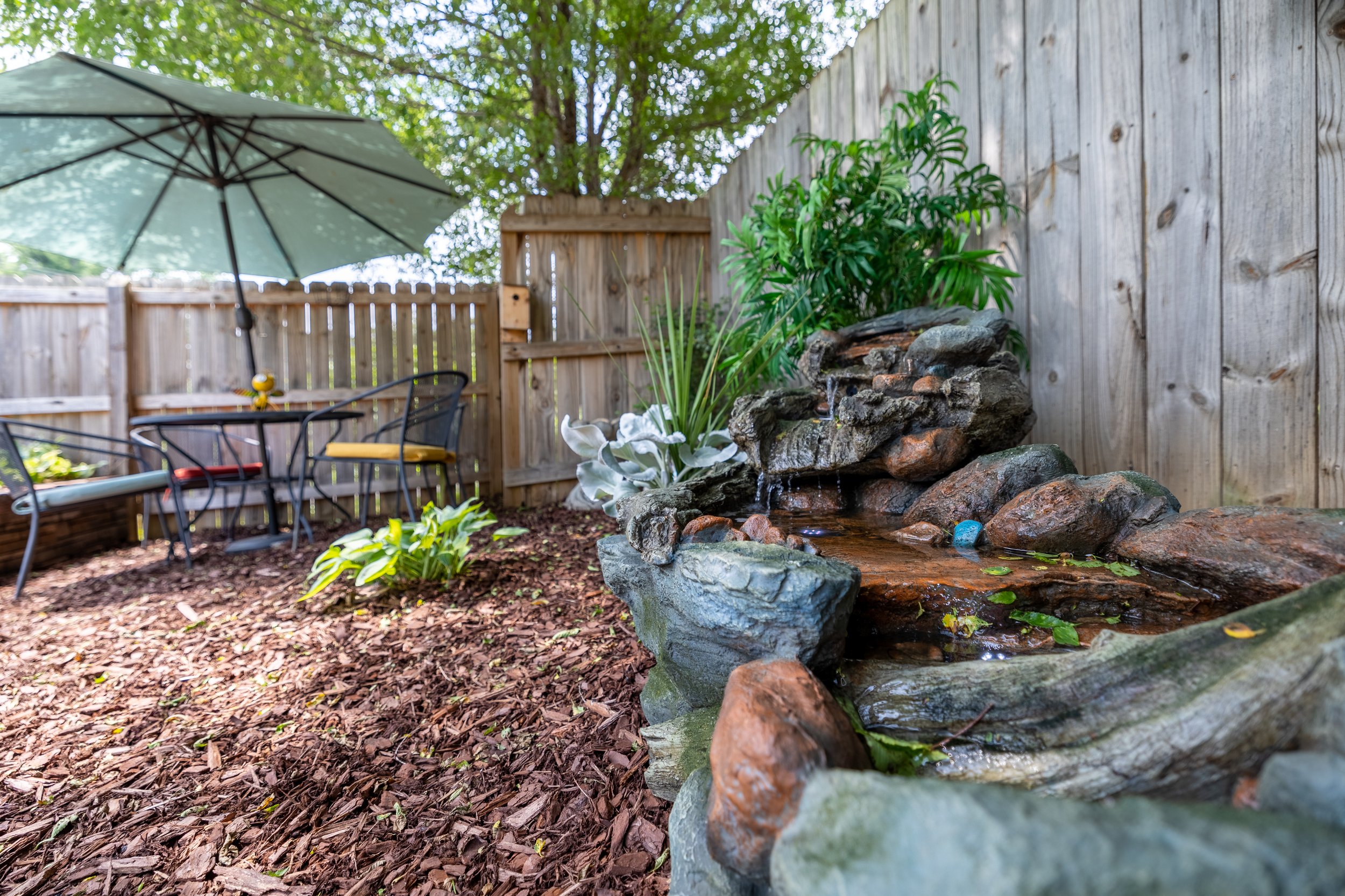 Backyard patio with a large umbrella, seating area, decorative waterfall feature, and plants enclosed by a wooden fence.