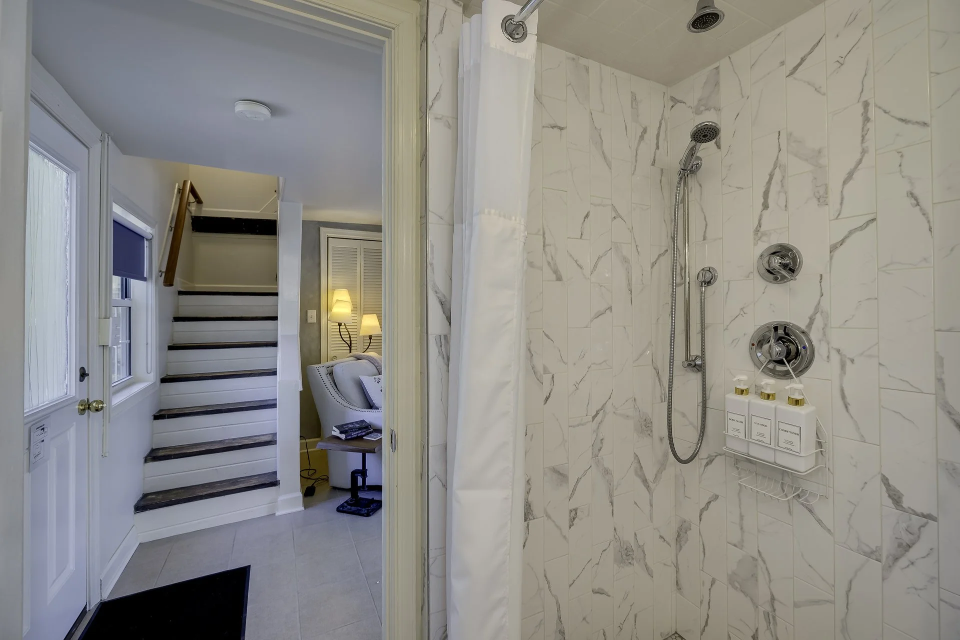 View of a marble tiled shower with multiple showerheads and a white shower curtain, next to a door opening to a living space with a staircase, windows, and a sitting area with lamps.