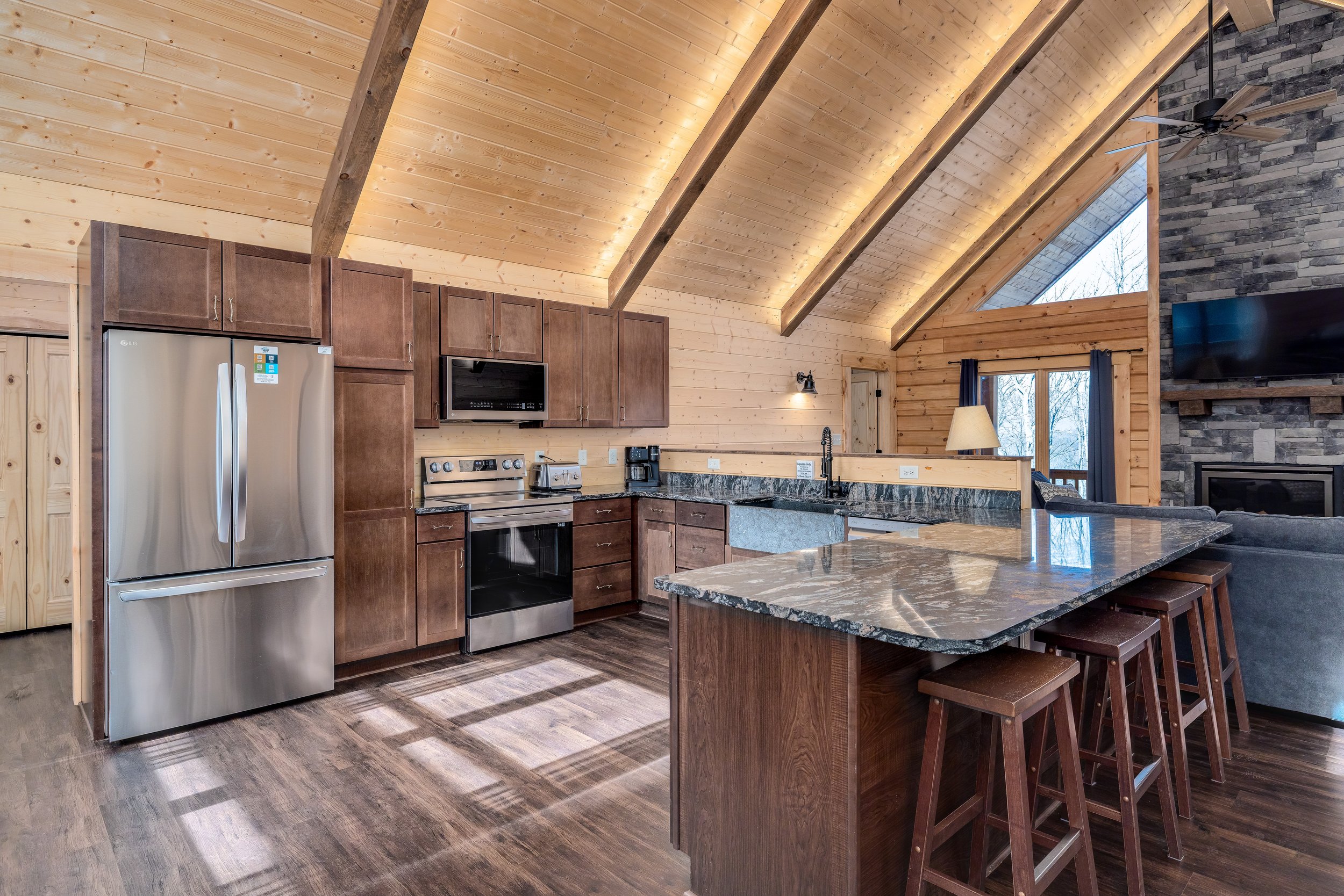Interior view of a modern kitchen with a vaulted wooden ceiling, stone fireplace, and large window with a view of trees. Features include stainless steel refrigerator, microwave, oven, granite countertops, and bar with stools.