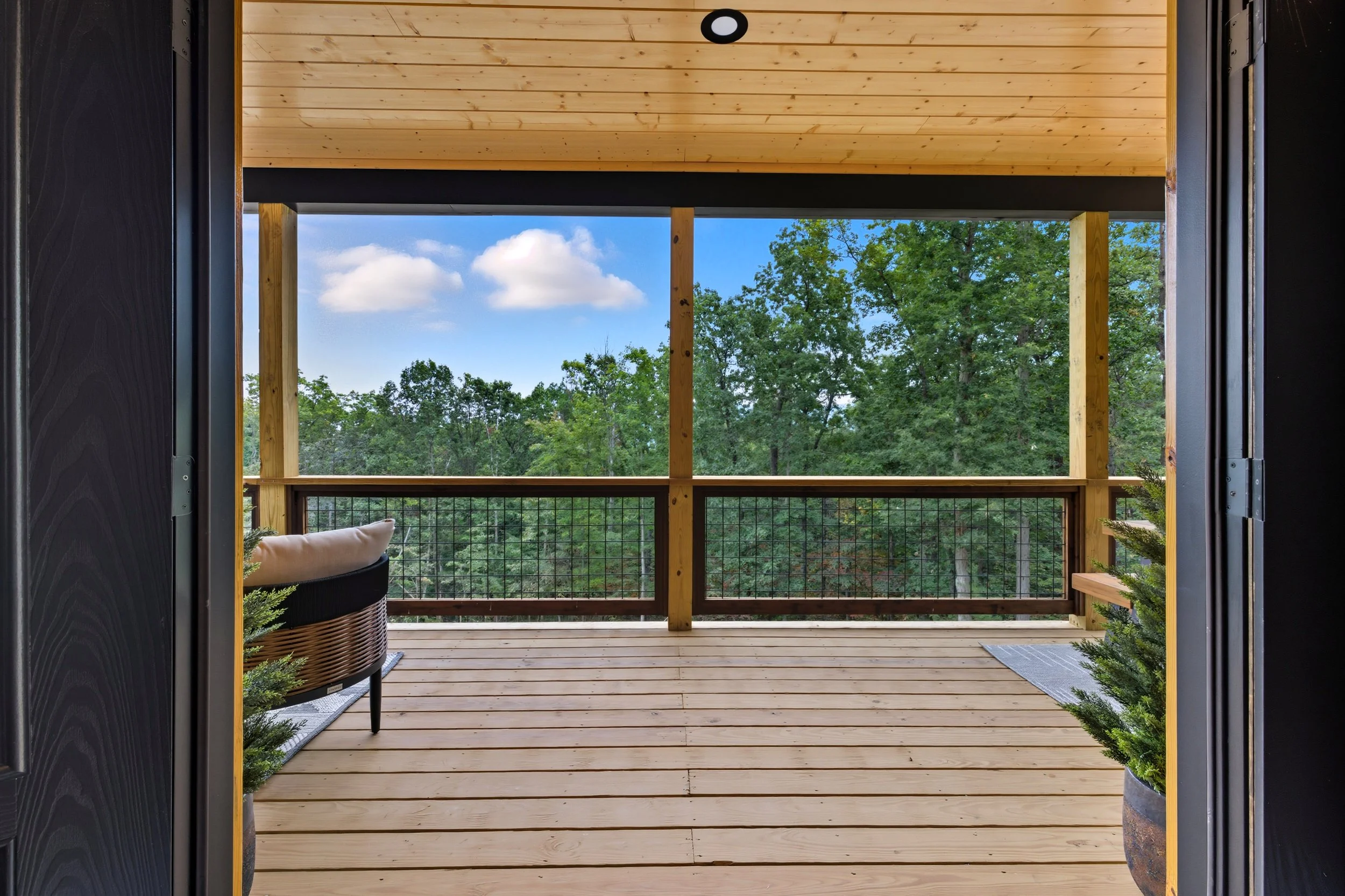 View from a doorway to a wooden balcony with a black railing, overlooking lush green trees and a partly cloudy sky.