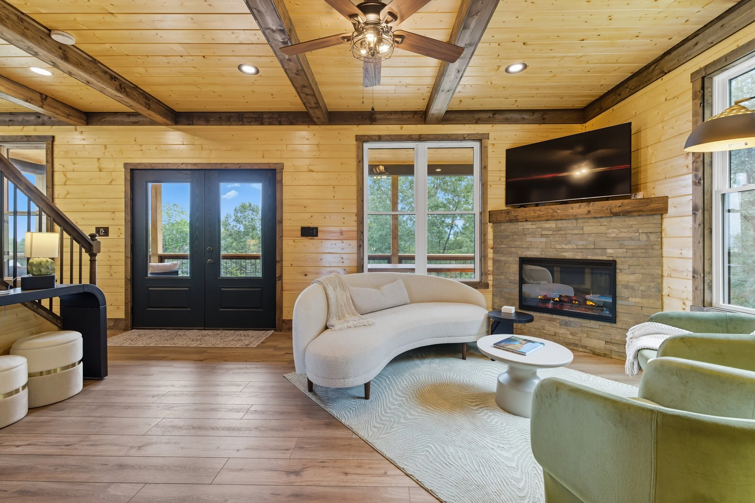 Living room with a wood-paneled ceiling, a ceiling fan, a stone fireplace with a TV mounted above it, and large windows showing trees outside. There is a cream-colored curved sofa with a blanket, a small black side table with magazines, and a lime-gr