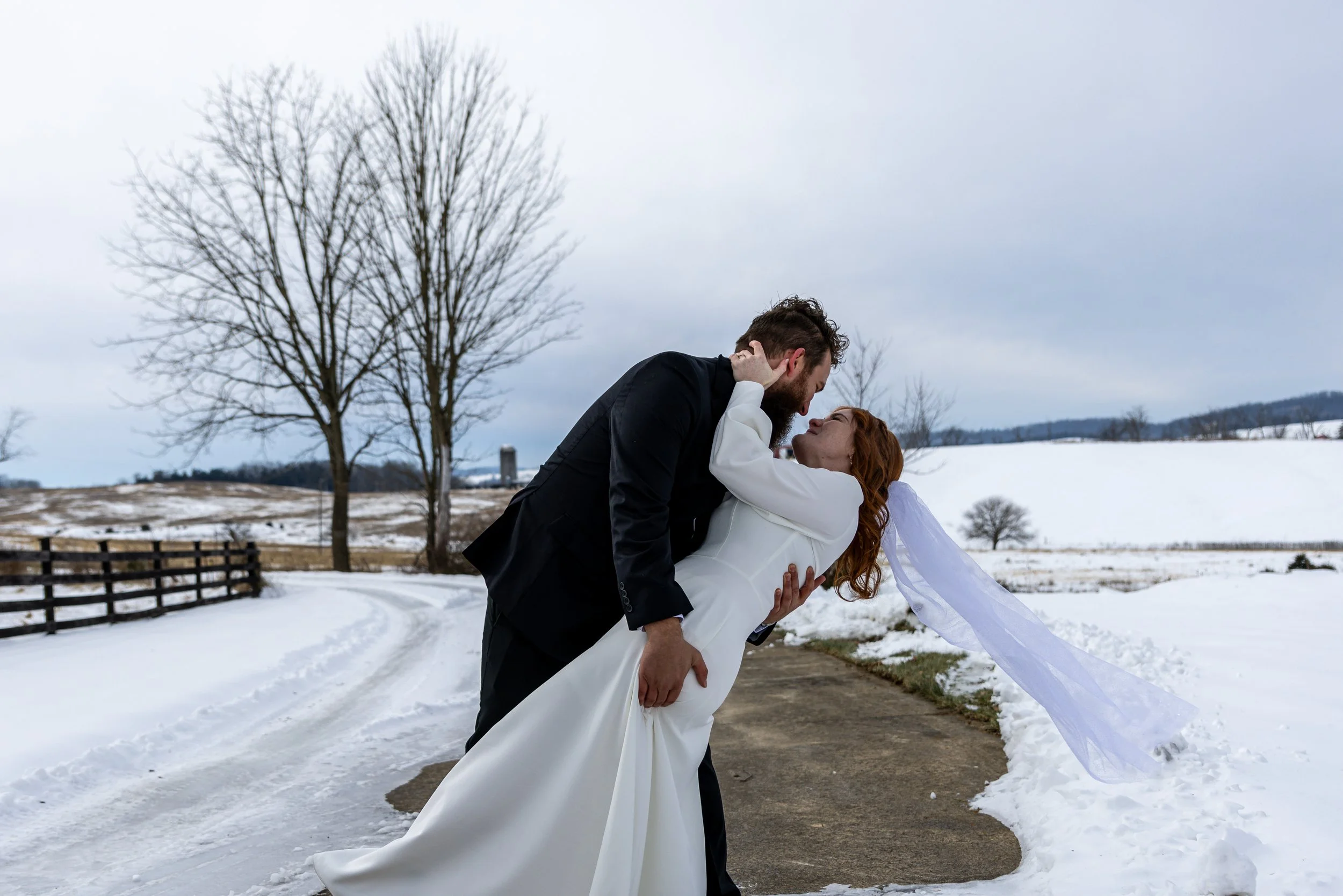A man dressed in a black suit is dipping a woman in a white wedding dress outdoors on a snowy day, with bare trees and a fence in the background.