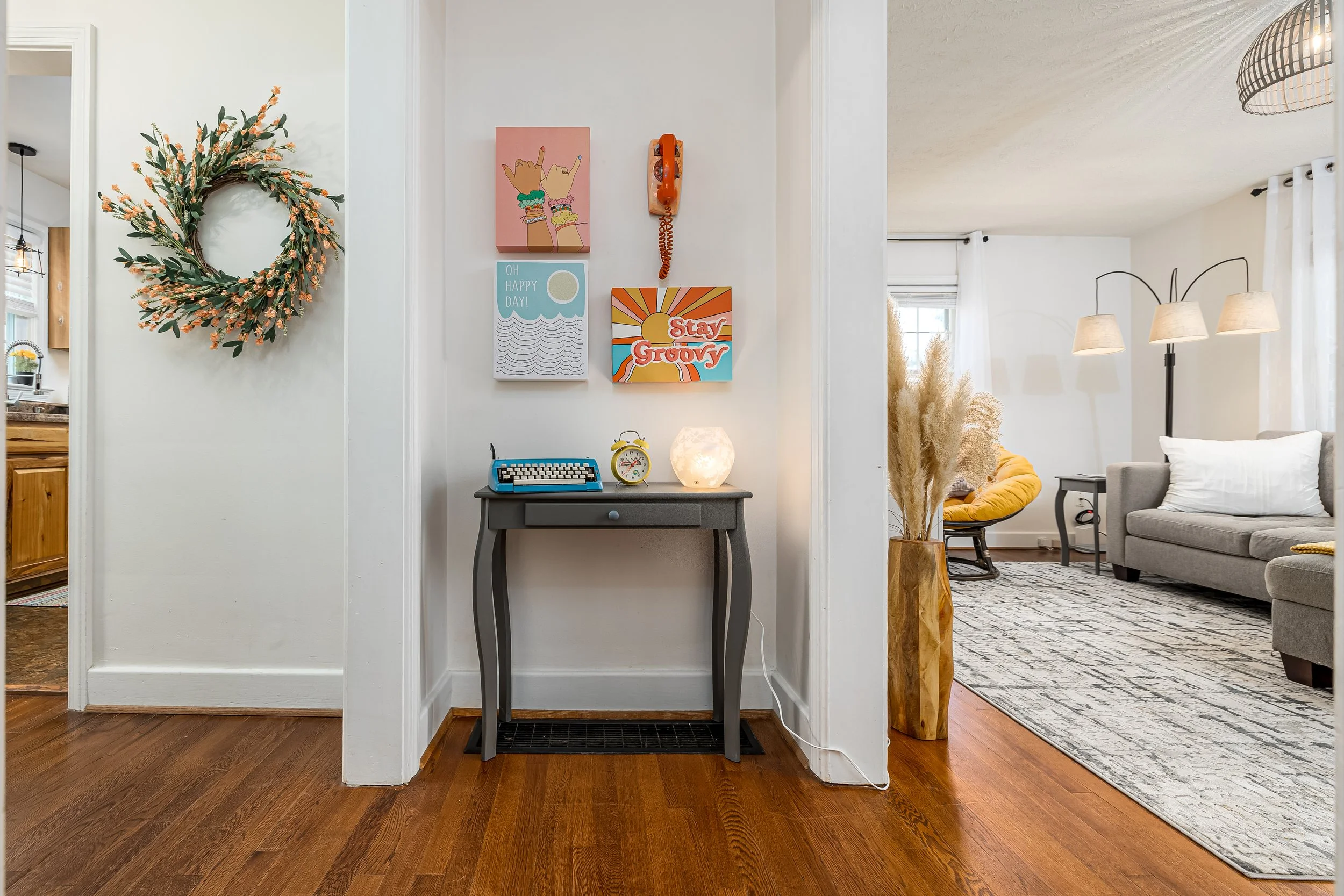 Living room with a hallway decorated with a wreath, colorful wall art, a vintage clock, a small table, and a lamp. The living room has a armchair, a side table, a floor lamp, and a rug.
