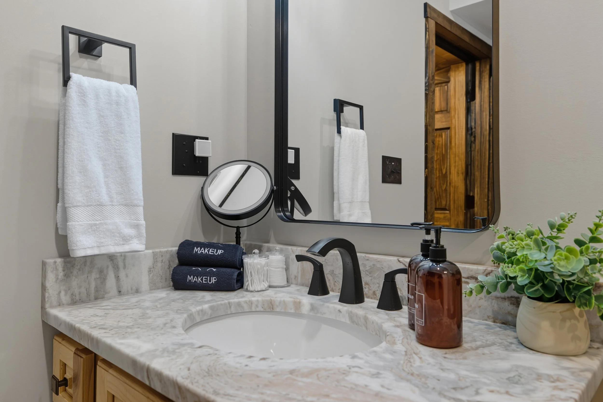 Bathroom vanity with a marble countertop, black faucet, and a large mirror. Items include folded towels labeled 'MAKEUP', a small mirror, a brown soap dispenser, a plant in a white pot, and various toiletries.