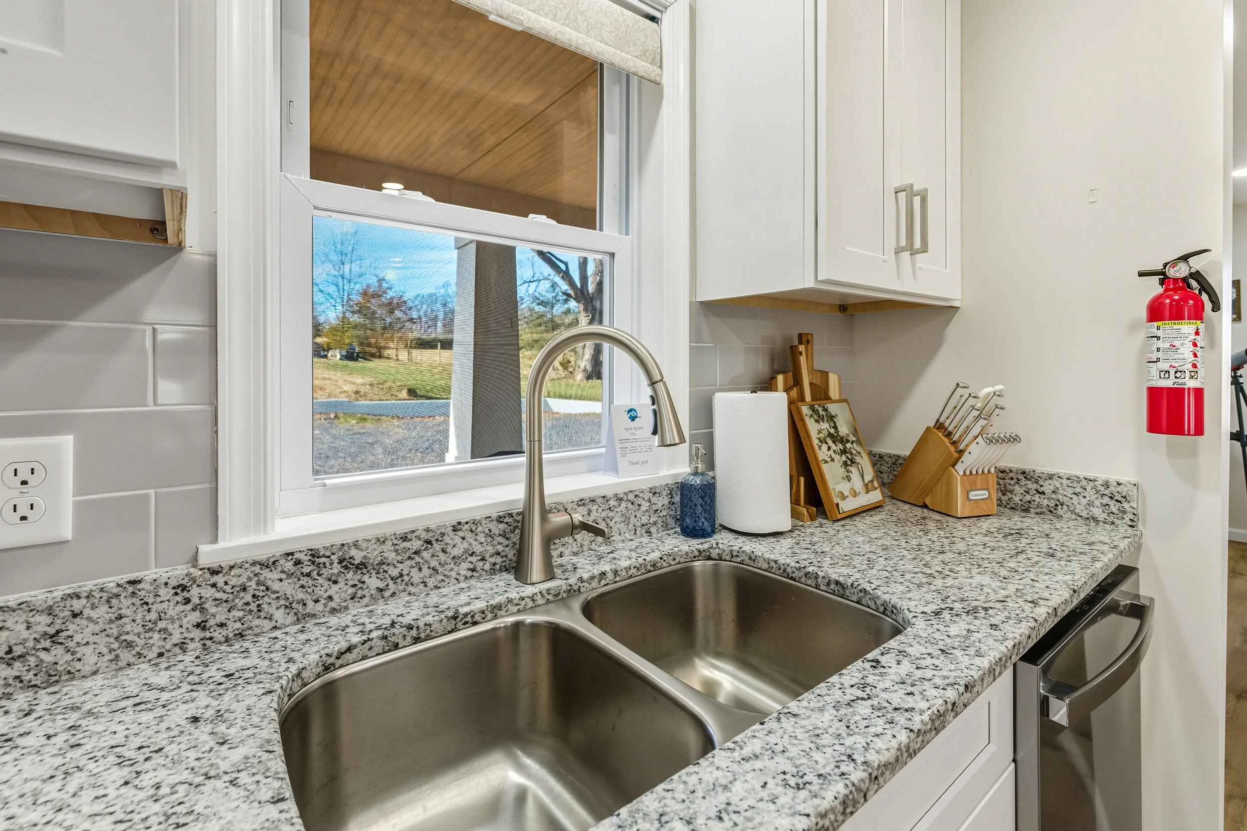 Kitchen counter with a granite surface, a stainless steel double sink, a faucet, a soap dispenser, a paper towel roll, a wooden knife block, and framed pictures, with a window overlooking an outdoor yard with trees.