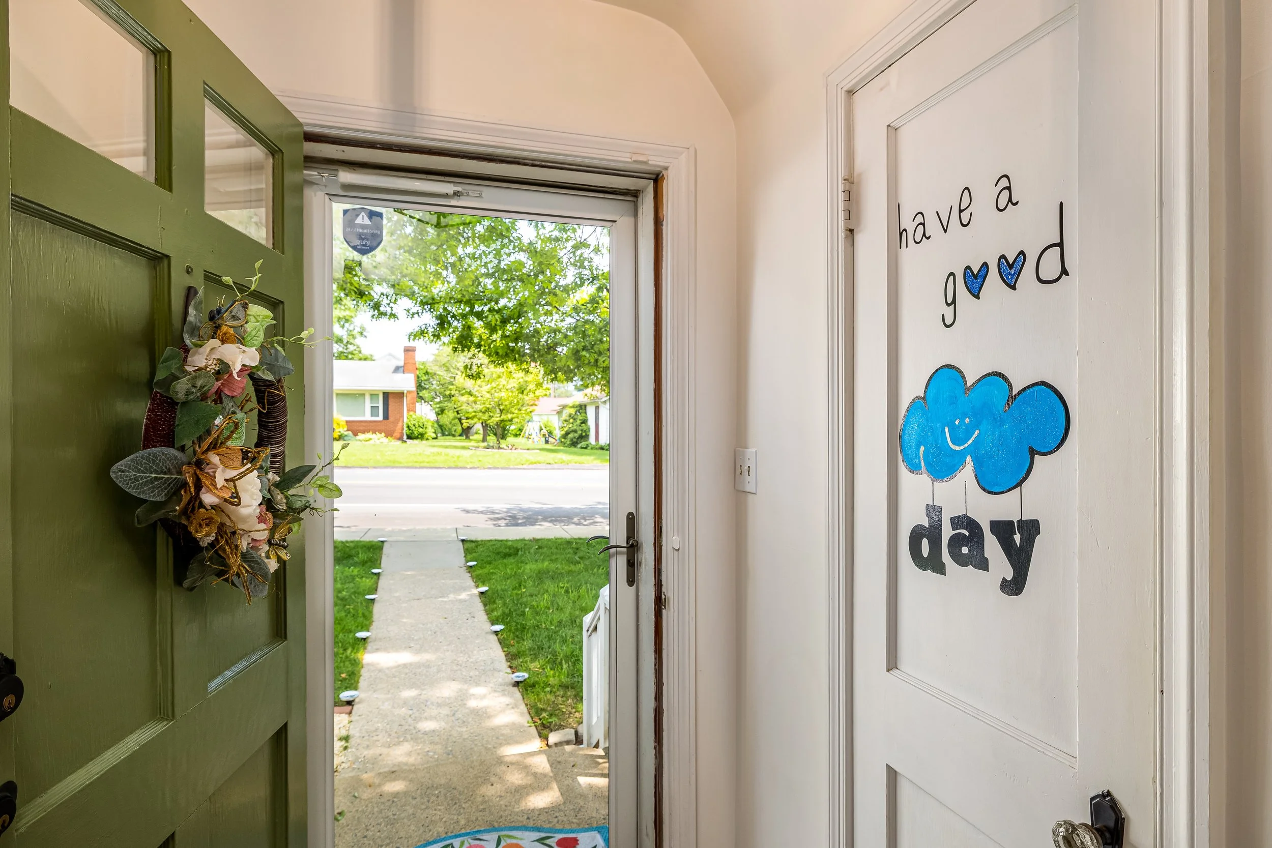 Door opens to a front yard with green trees and houses in the background, inside the house, a wall with the words "have a good day" and a blue smiling cloud decoration.