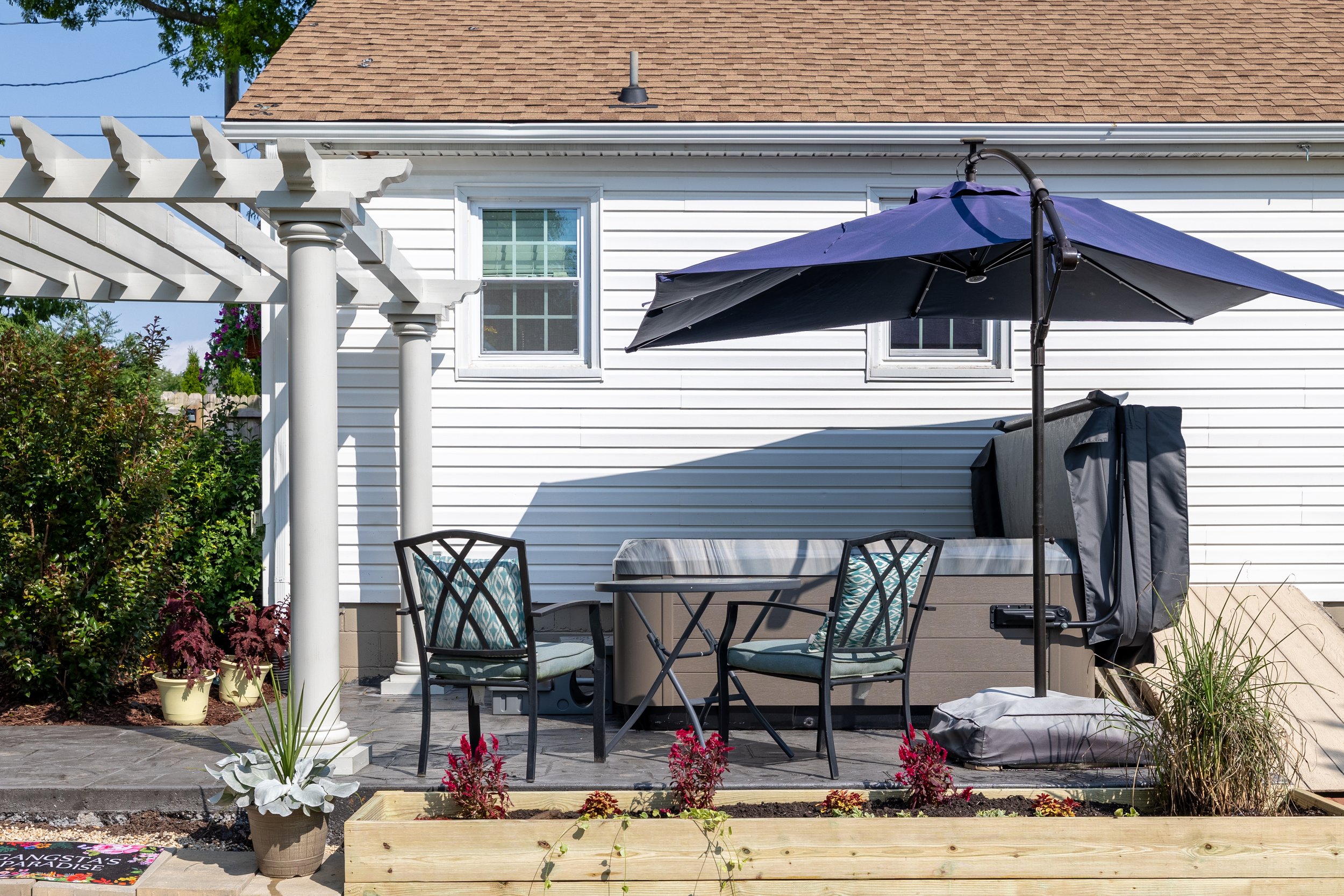 Backyard patio with outdoor dining set, large blue umbrella, potted plants, and garden bed with flowers and vegetables, in front of white house with brown roof and two windows.